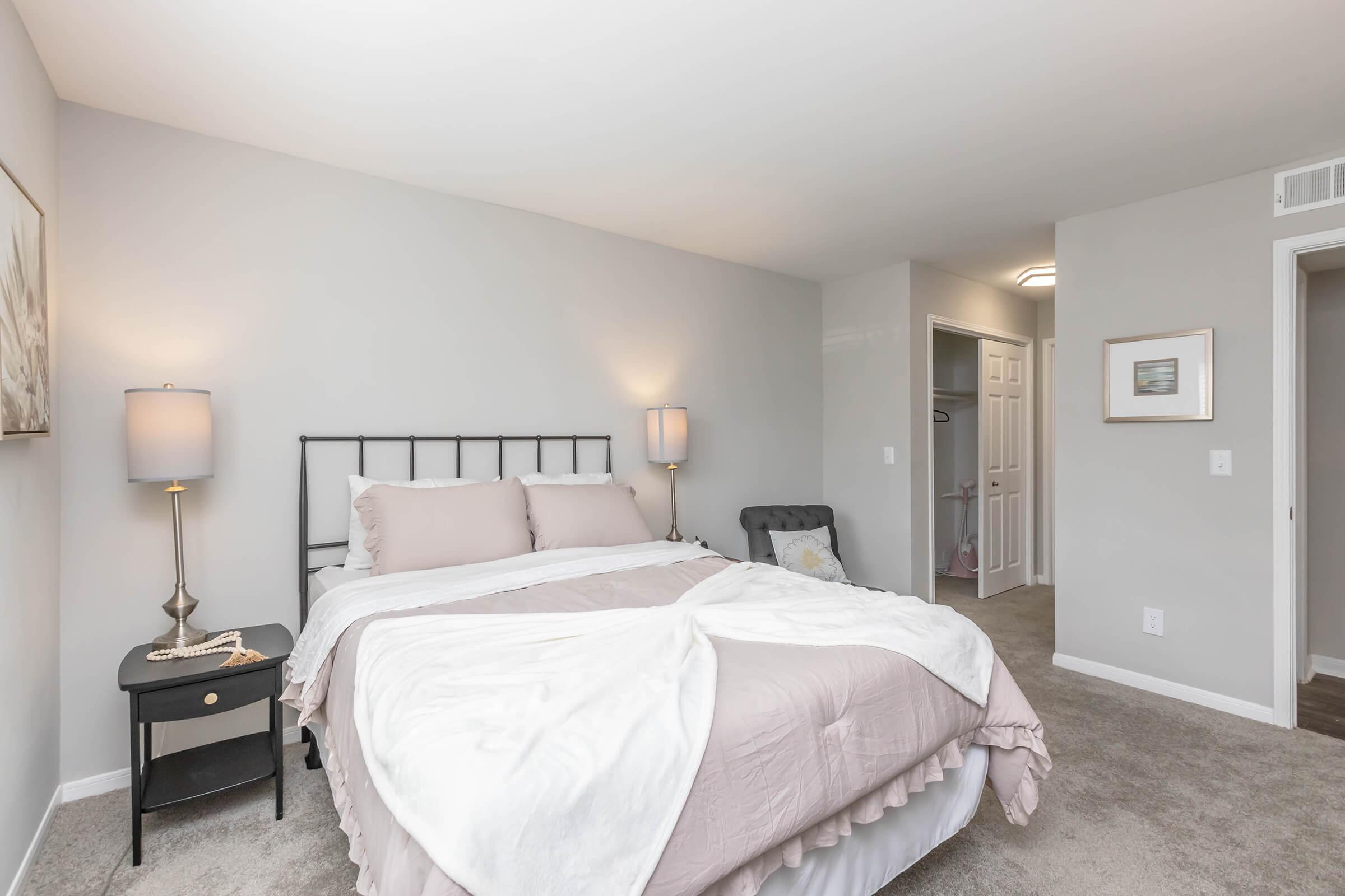 A modern bedroom featuring a light gray color scheme. The bed has neutral bedding and decorative pillows. A bedside lamp and a small side table are visible next to the bed. A closet is partially open in the background, and the walls are decorated with simple art. The room has a cozy and inviting atmosphere.