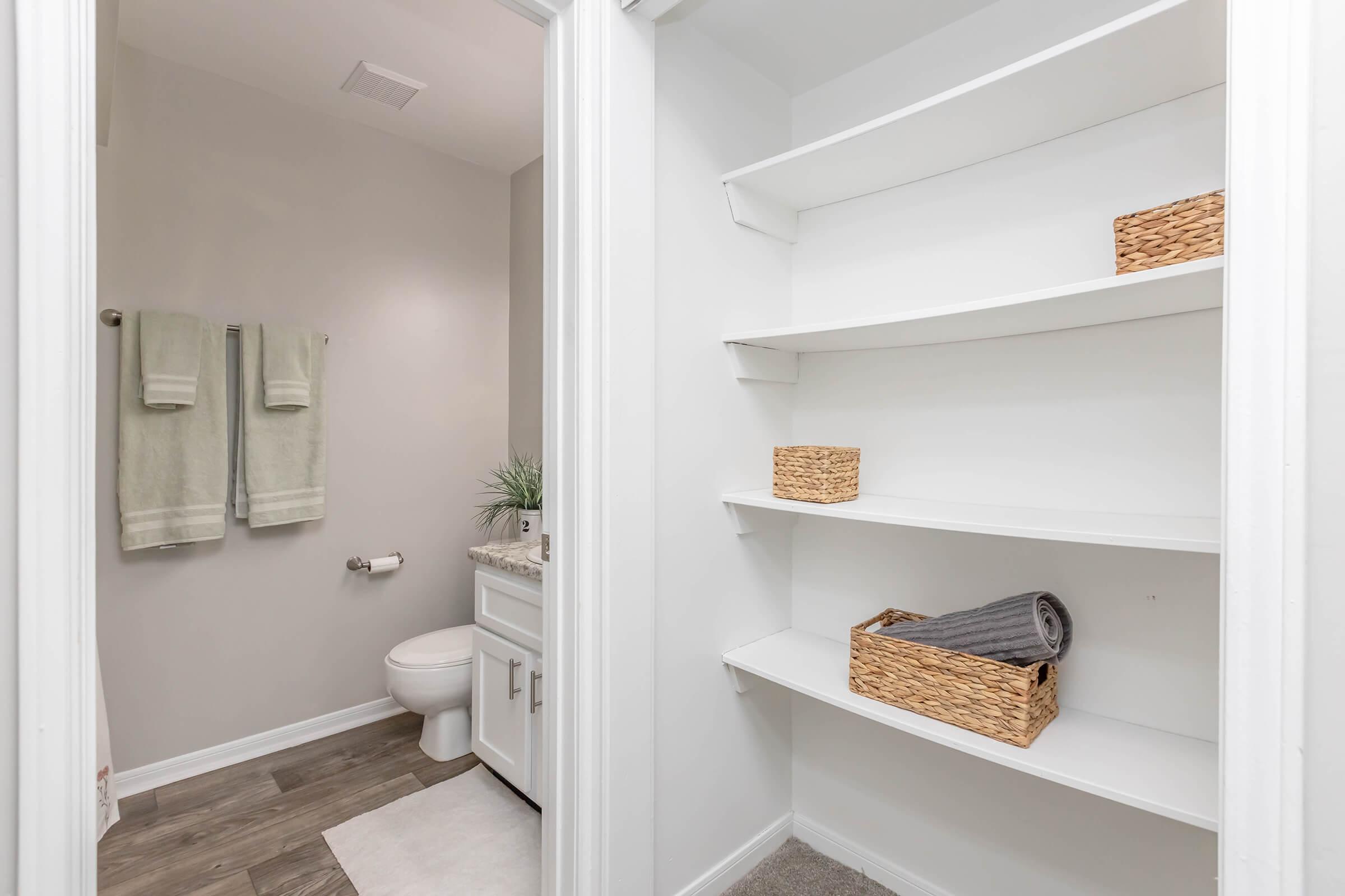 A well-organized bathroom featuring a toilet, a sink with a countertop, and two green towels hanging on the wall. There are three open shelves with woven baskets and a rolled towel, set against a light gray wall and wooden floor. Bright and clean environment.