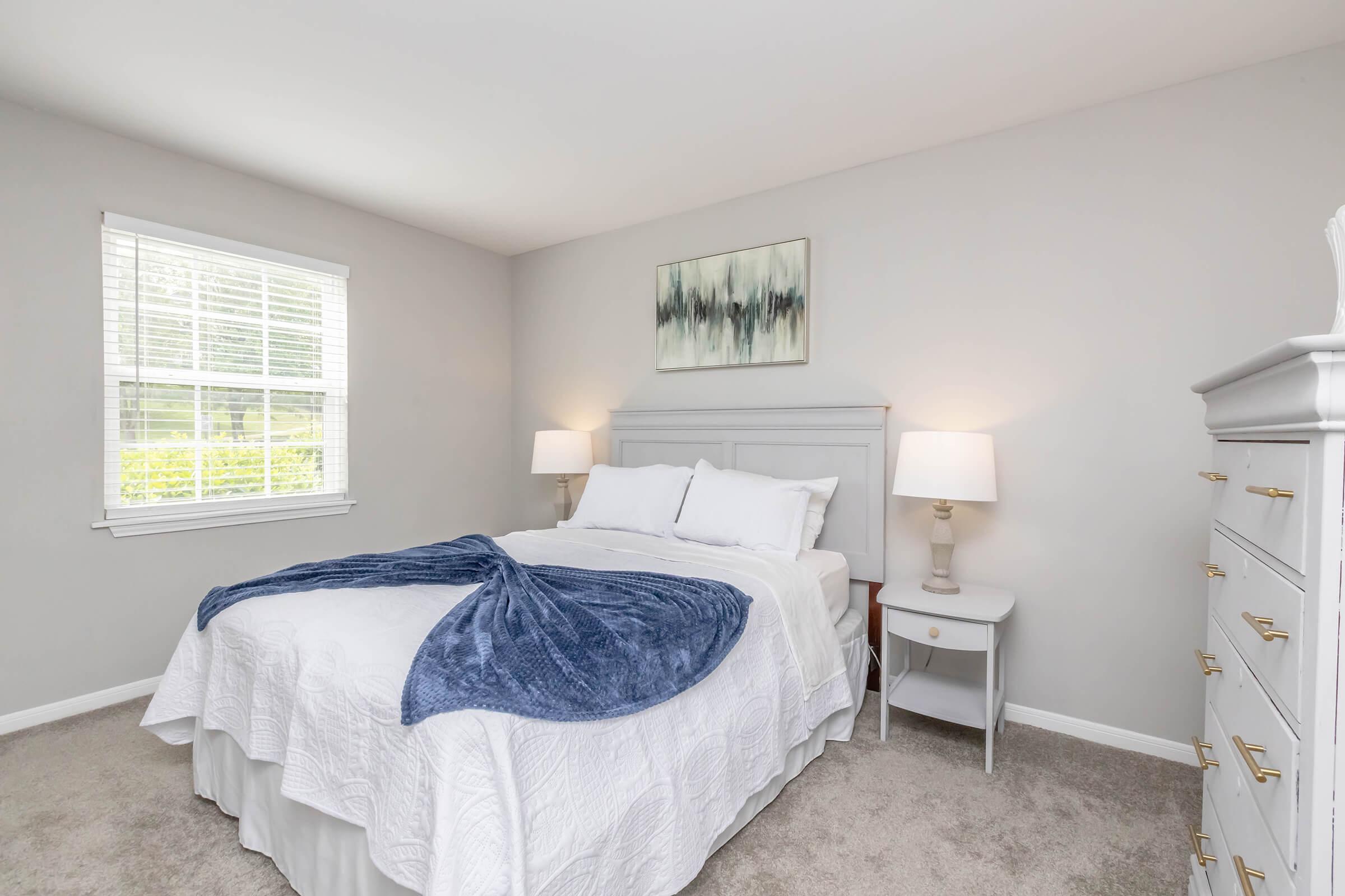A cozy, well-lit bedroom featuring a white bedspread with a blue throw blanket, two bedside lamps, and a white nightstand. The room has light gray walls, a large window with white blinds, and an abstract painting above the headboard, creating a peaceful atmosphere.