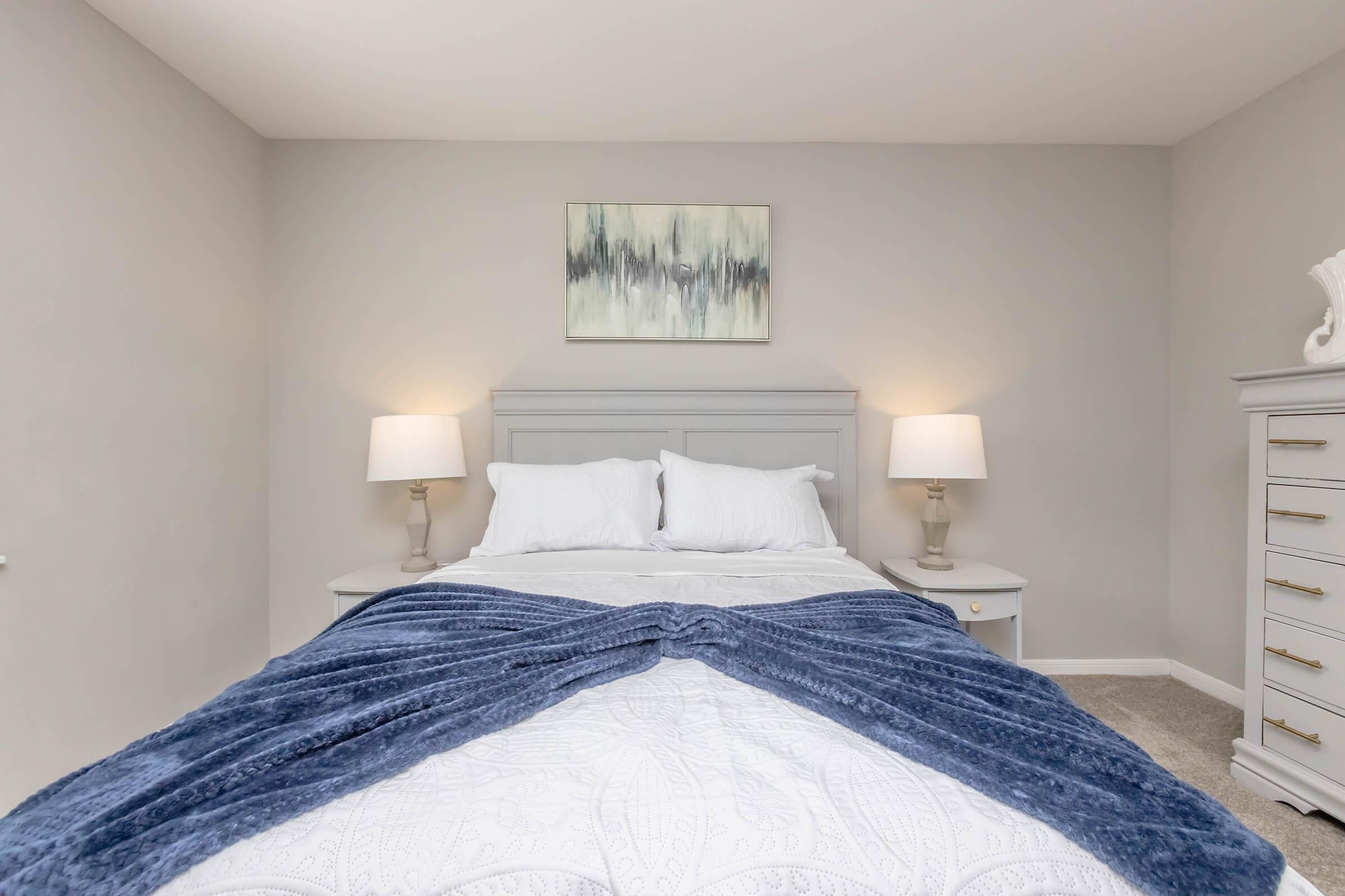 A neatly arranged bedroom featuring a light gray wall, a queen-sized bed with white linens and a blue throw blanket. Two bedside tables with lamps flank the bed, and a decorative piece hangs above. A light gray dresser is positioned against the wall, adding to the cozy and modern aesthetic.