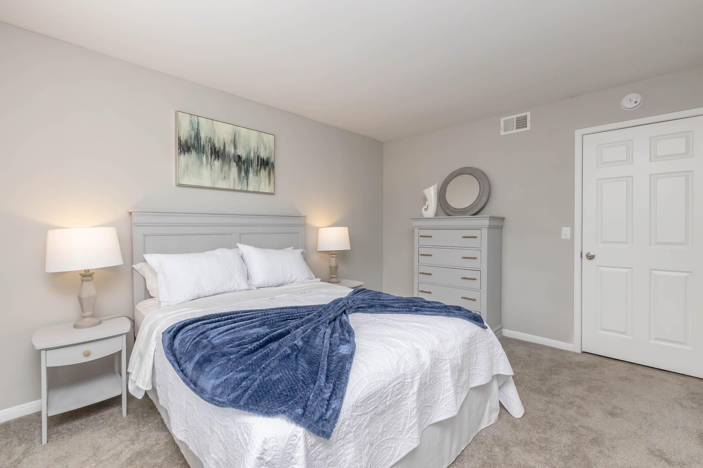 A cozy and well-decorated bedroom featuring a queen-sized bed with white bedding and a navy blue throw blanket. There are two bedside lamps on small tables, a dresser, and a round mirror on the wall. The walls are painted in a soft gray tone, creating a calm atmosphere.