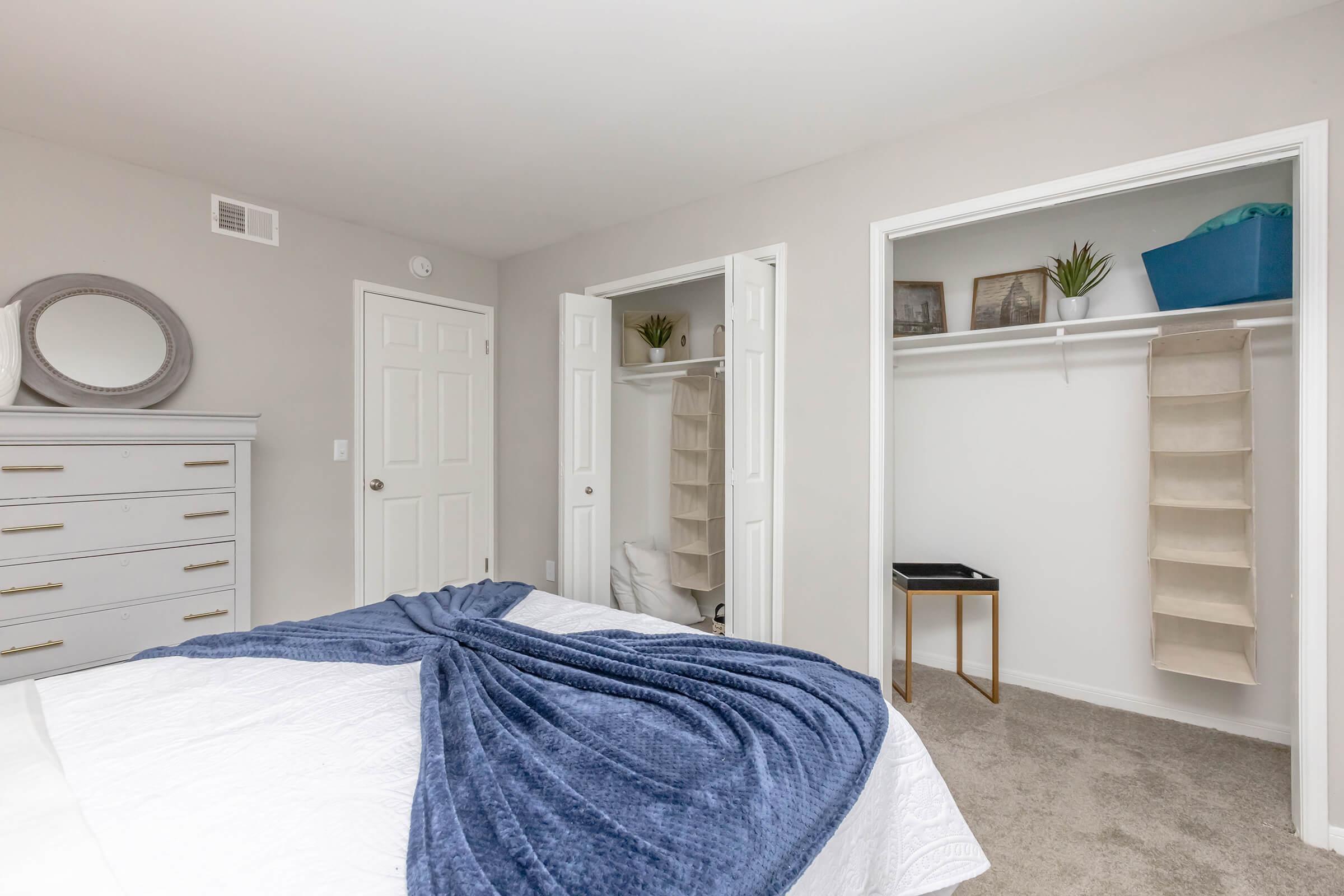 A neatly designed bedroom featuring a bed with a blue throw blanket, a dresser with a round mirror above it, and two closets with open doors. One closet has shelves, while the other is partially empty. The walls are painted light gray, and the carpet is a soft beige color.