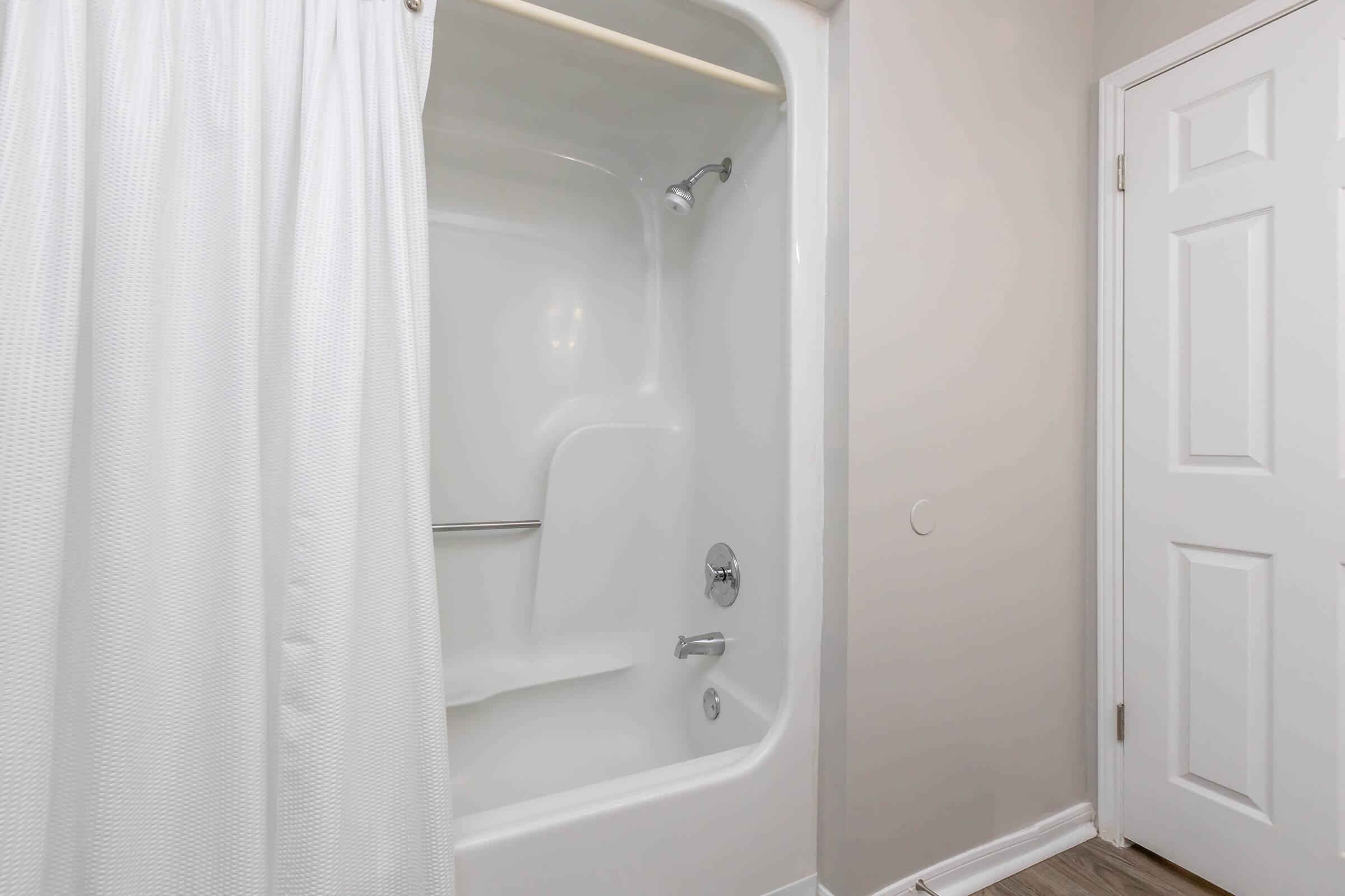 A clean, modern bathroom featuring a shower-tub combo with a white curtain. The walls are painted in a light color, complementing the neutral flooring. A closed door is visible on the right side of the image, adding to the overall spacious feel of the room.