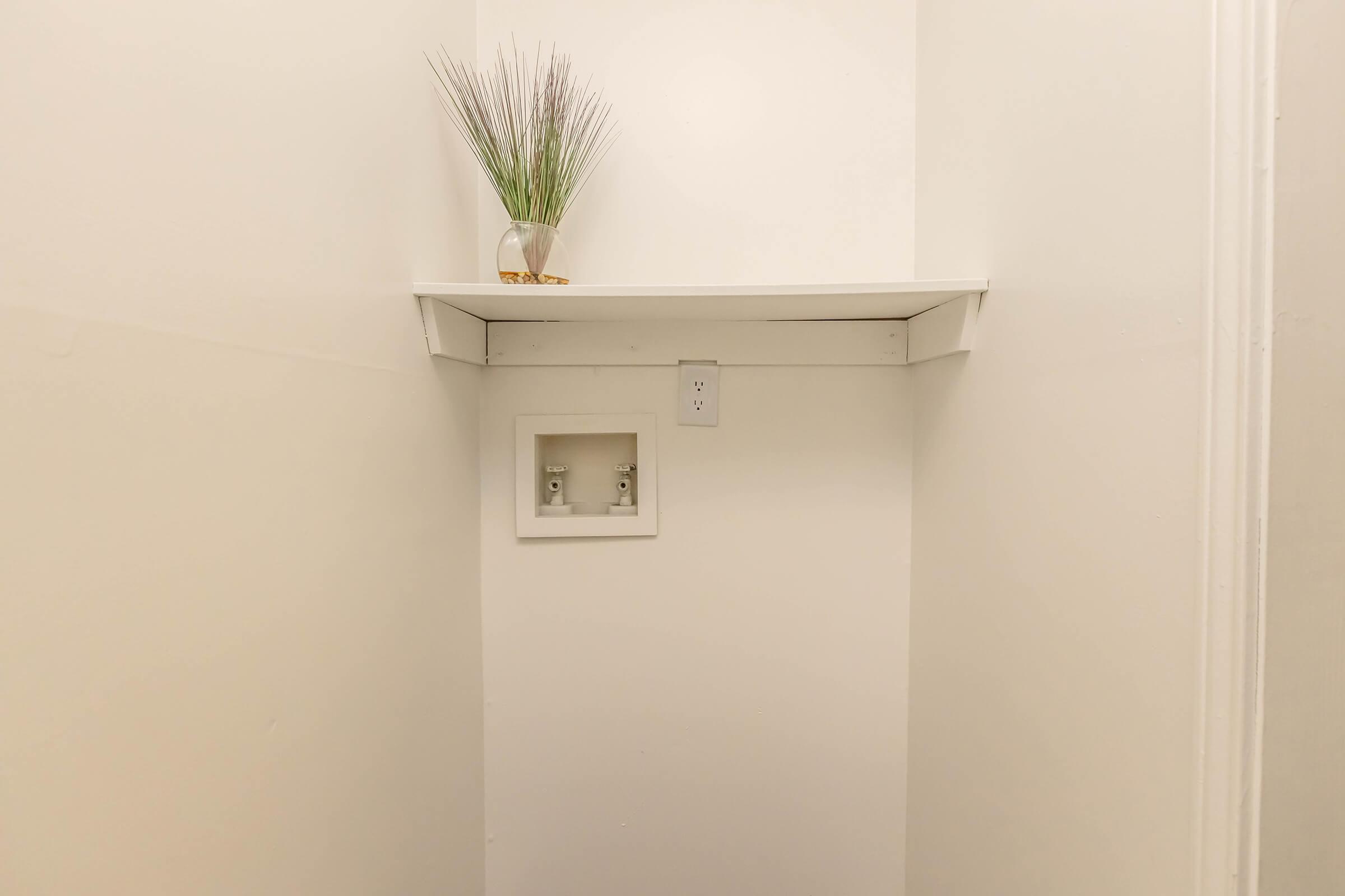 A minimalistic corner of a room featuring a white wall. There is a small white shelf mounted near the top with a decorative vase containing greenery. Below the shelf, a white inset space with two electrical outlets is visible. The overall appearance is clean and simple.
