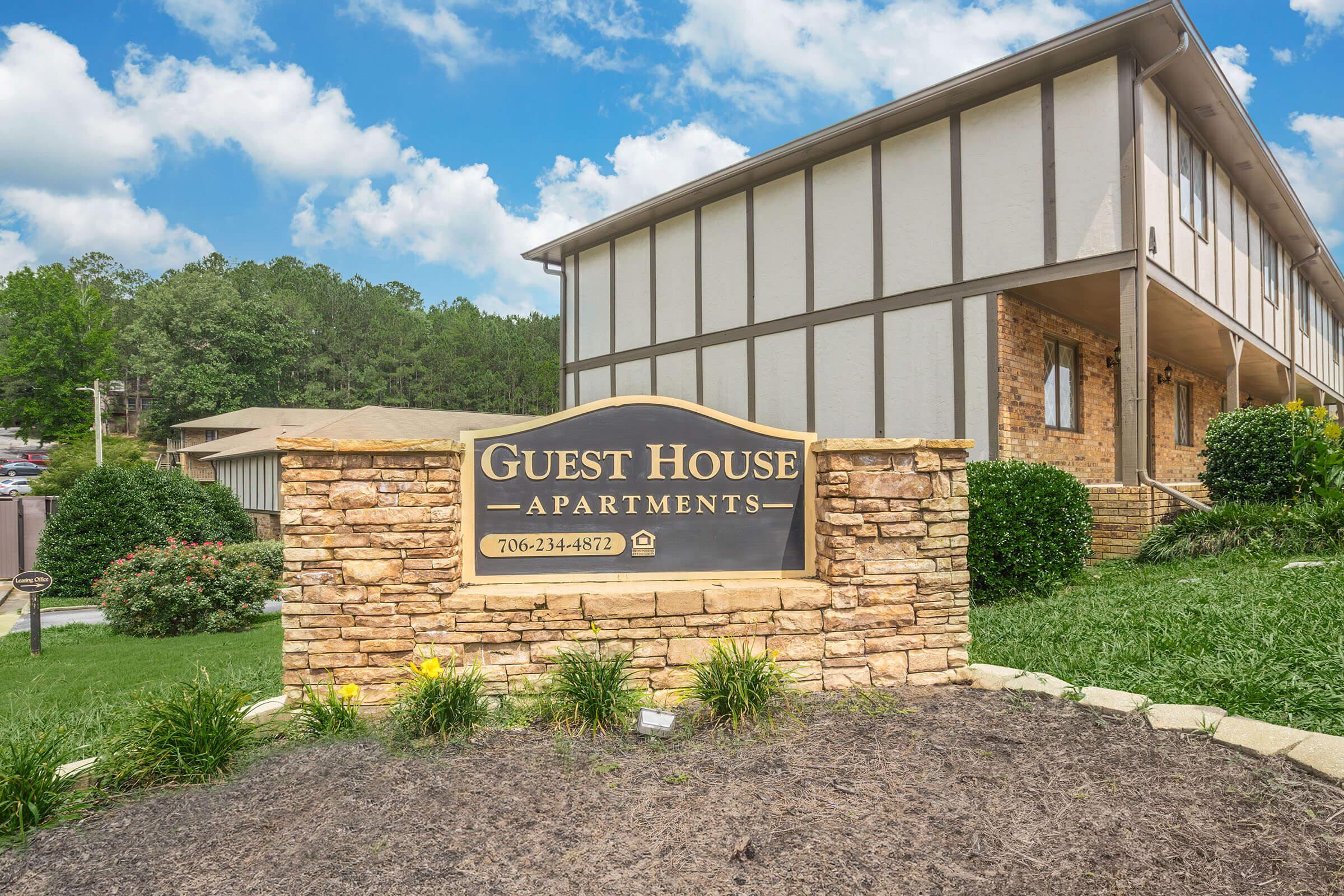 Sign for Guest House Apartments made of stone, featuring the text "Guest House Apartments" prominently. The building is two stories with a tan exterior, surrounded by green grass and landscaped bushes. The sky is partly cloudy, creating a welcoming atmosphere.