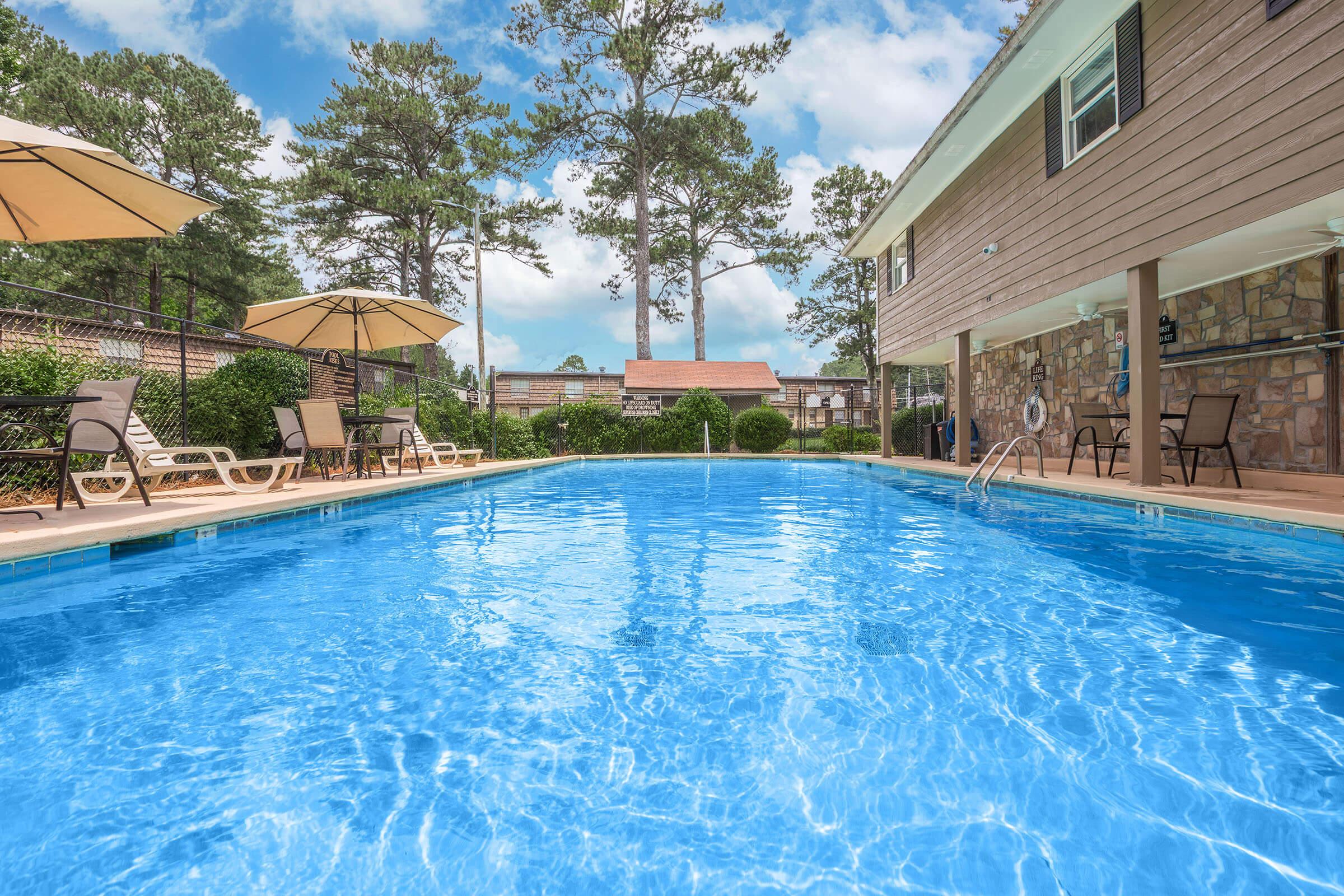 A clear blue swimming pool surrounded by lounge chairs and umbrellas, set in a landscaped area with trees in the background. A two-story building with a stone façade is partially visible, contributing to a serene outdoor atmosphere. The scene is bright and inviting, perfect for relaxation and enjoyment by the pool.