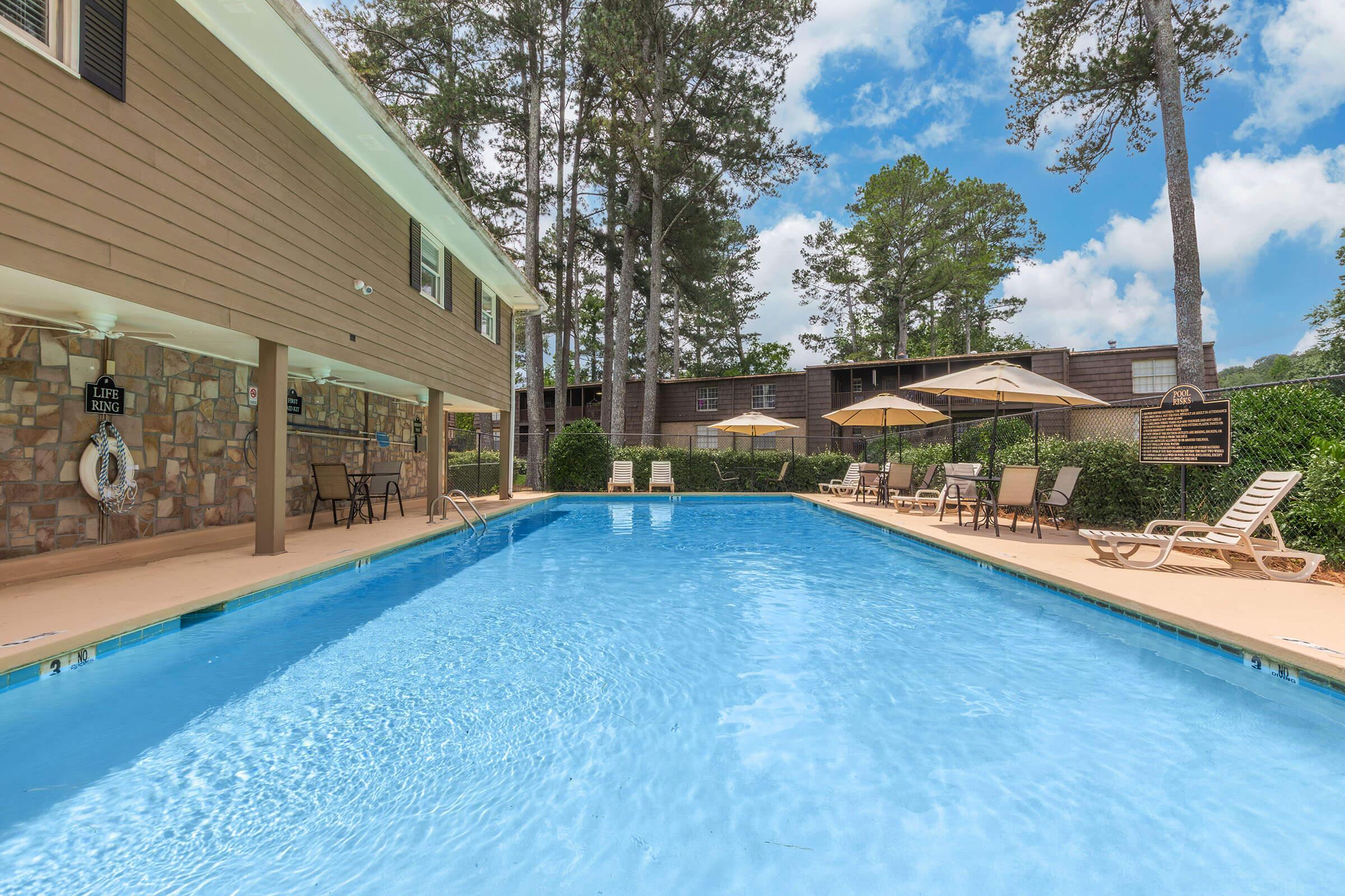 A clear blue swimming pool surrounded by sun loungers and umbrellas, with a stone wall on one side and trees in the background. The sky is bright with fluffy clouds, creating a serene and inviting atmosphere for relaxation.