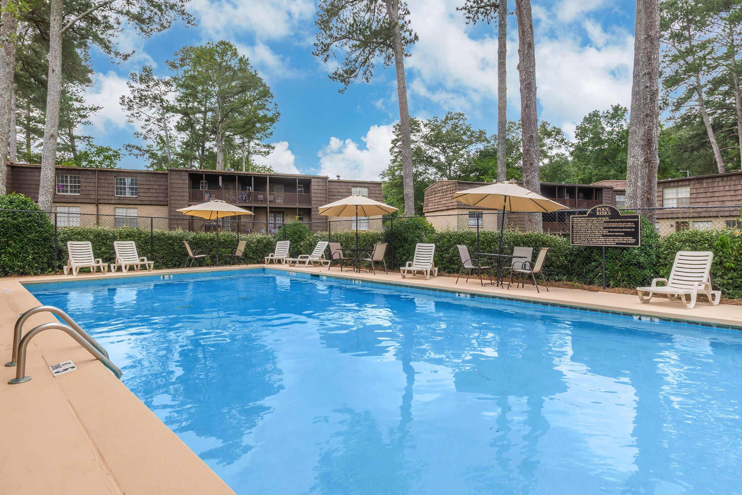 A clear swimming pool surrounded by lounge chairs and umbrellas, with green hedges and trees in the background. Apartment buildings are visible in the distance, under a partly cloudy blue sky. The scene conveys a relaxing outdoor atmosphere suitable for leisure.