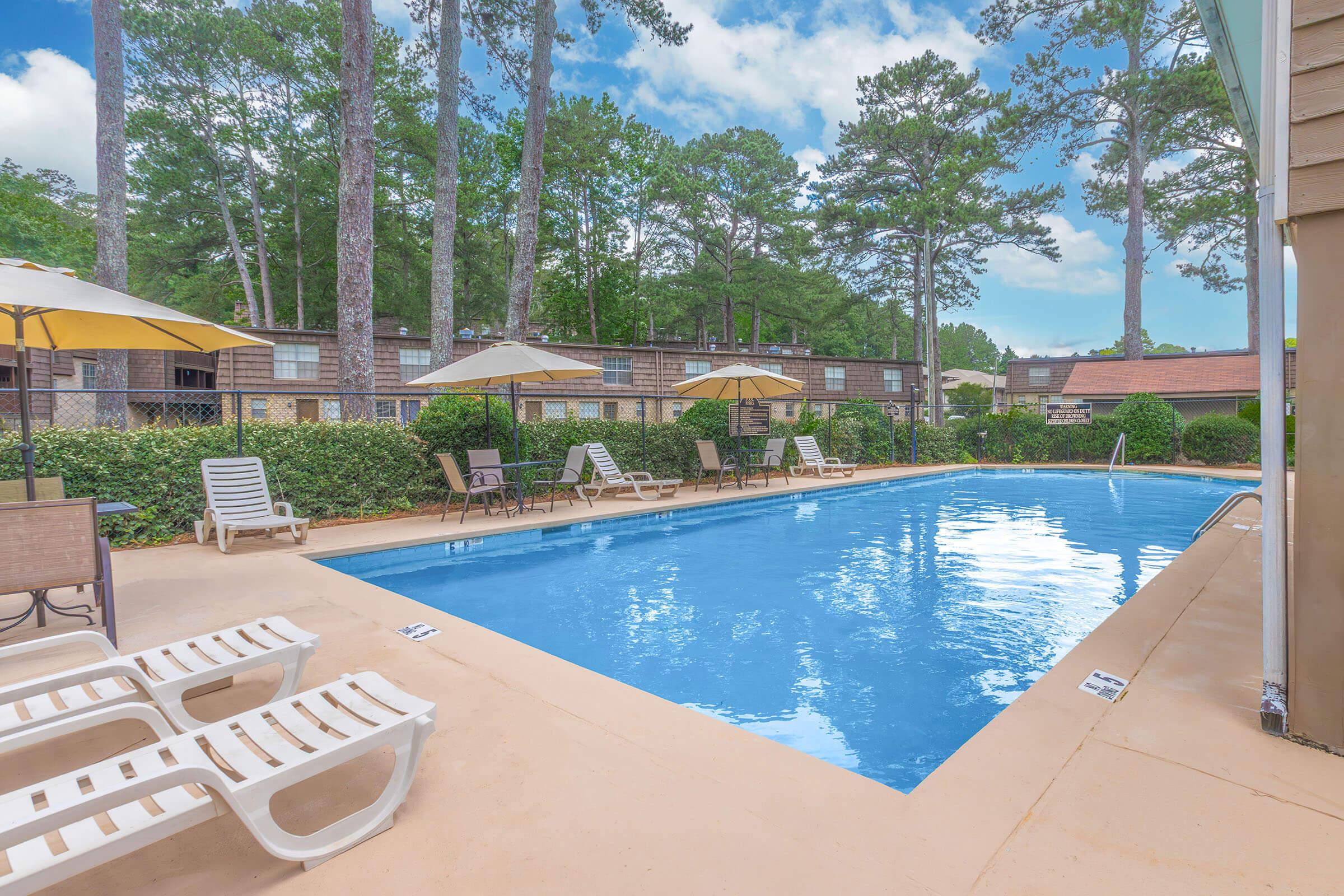 A sunny outdoor pool area featuring a clear blue pool surrounded by lounge chairs and umbrellas. Green trees and bushes provide a natural backdrop, while nearby buildings are visible in the background. The scene is inviting and perfect for relaxation on a warm day.
