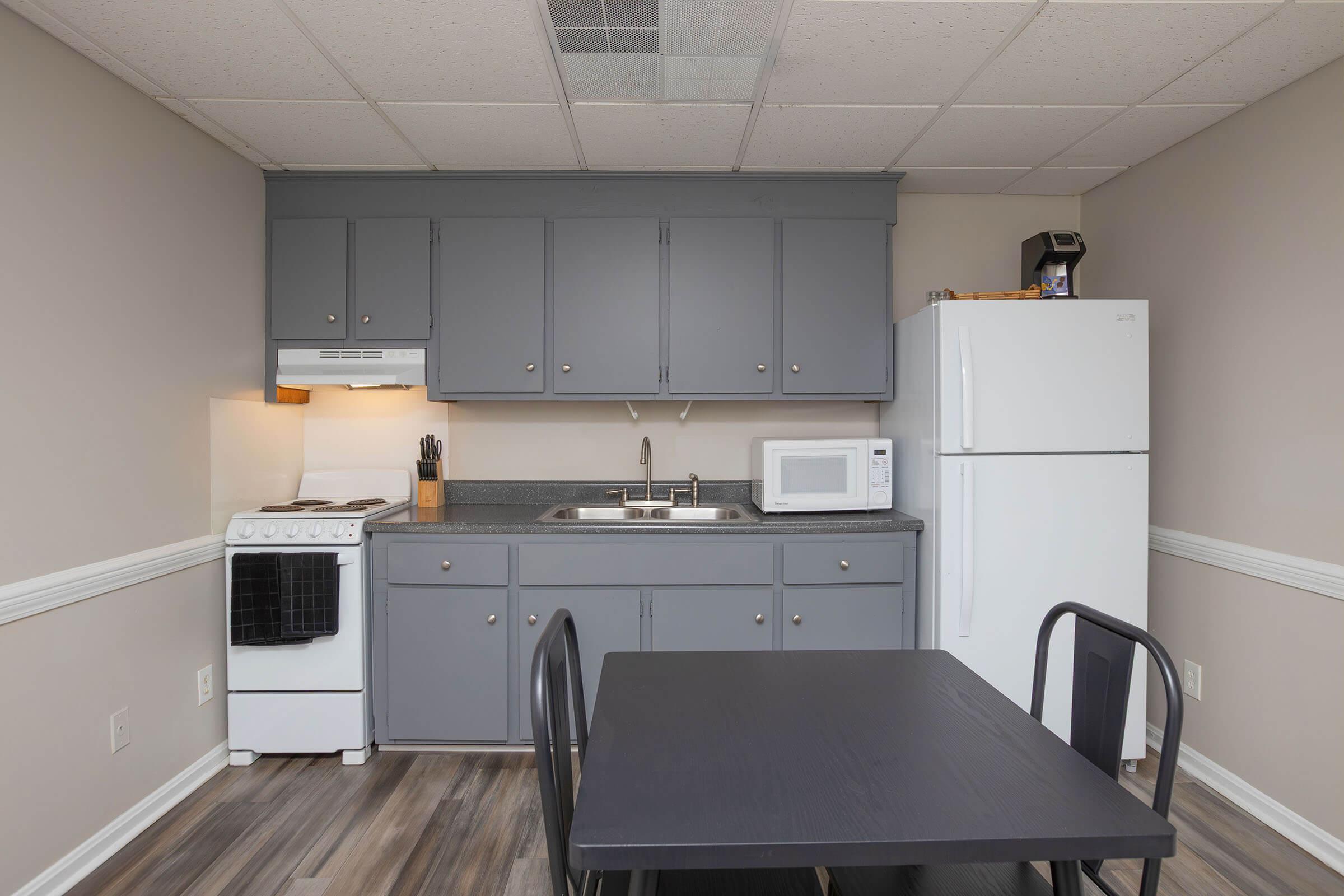 A modern kitchen with gray cabinets, a stove, sink, and microwave on the countertop. A white refrigerator stands next to the cabinets. There's a black dining table with two chairs in the foreground, and the floor features a wood-like laminate. The walls are painted a neutral color, enhancing the bright atmosphere.