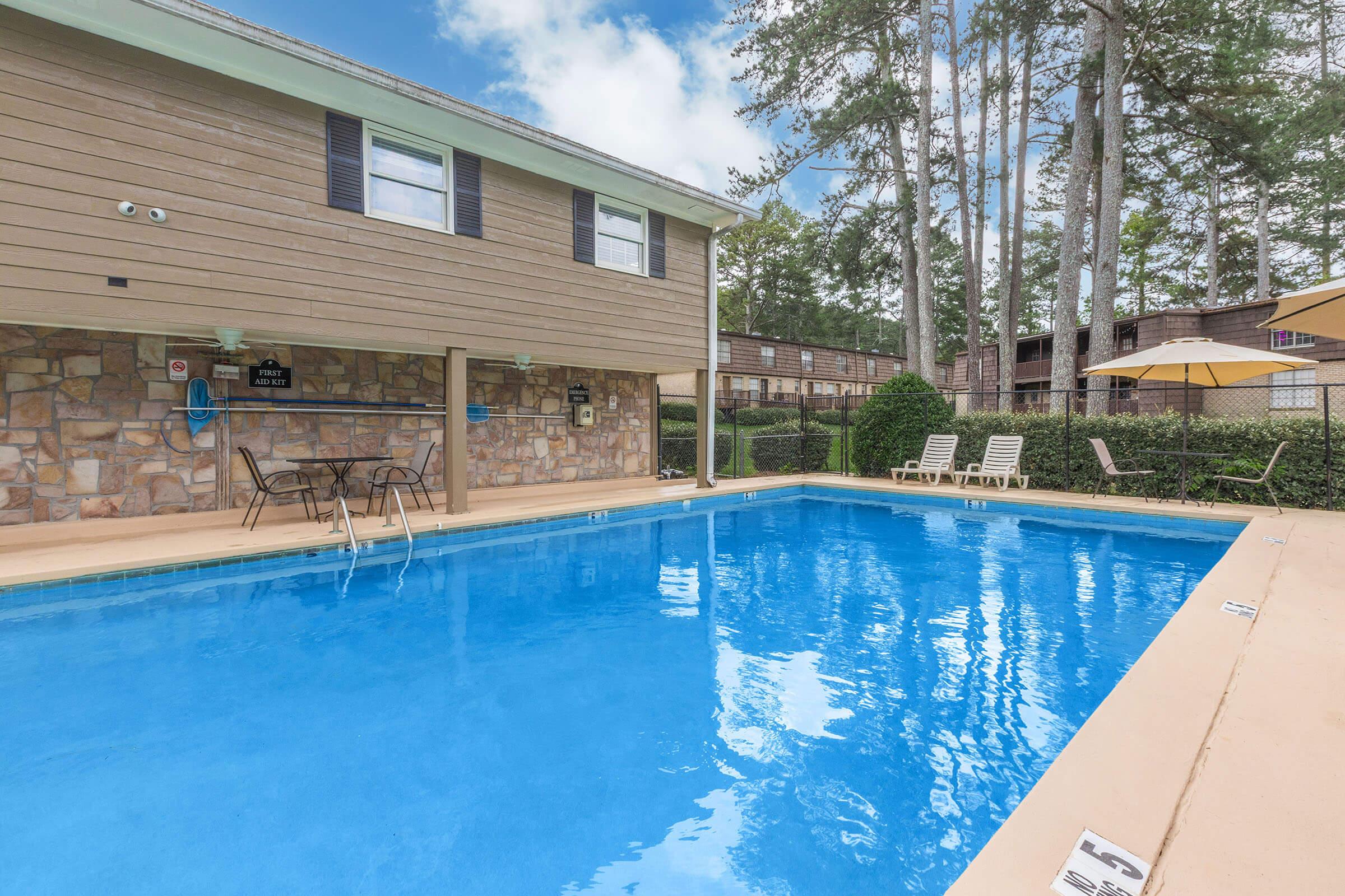 A crystal-clear swimming pool surrounded by a fenced area and tall trees. In the foreground, there are lounge chairs positioned near the water, and a patio set with chairs and a table. The building in the background has a rustic design, featuring wooden siding and windows. A sunny sky adds to the inviting atmosphere.