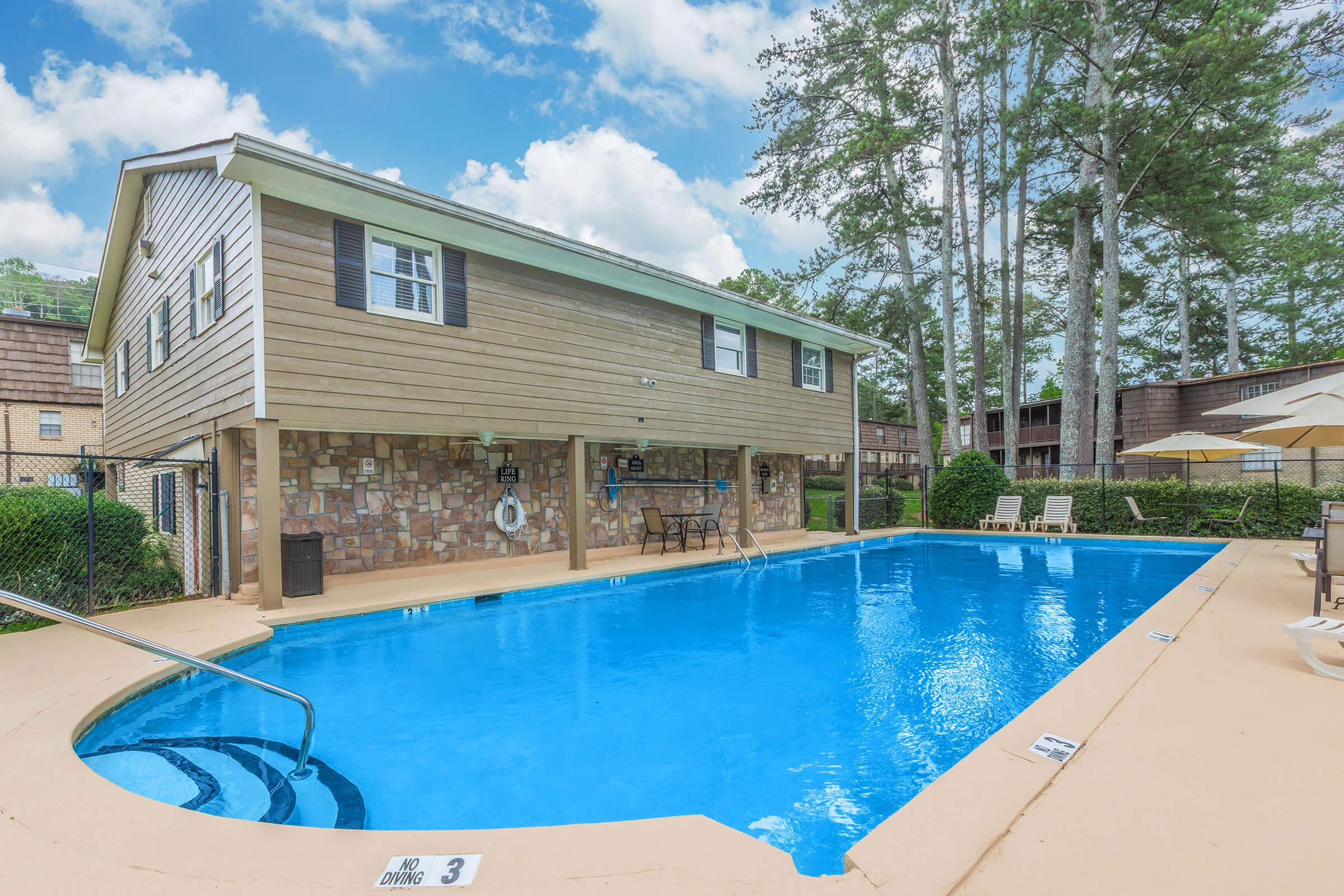 A residential pool area featuring a blue swimming pool surrounded by a patio. There are lounge chairs, a table with chairs, and a two-story building in the background. Lush green trees and a cloudy sky can be seen, creating a serene outdoor setting.