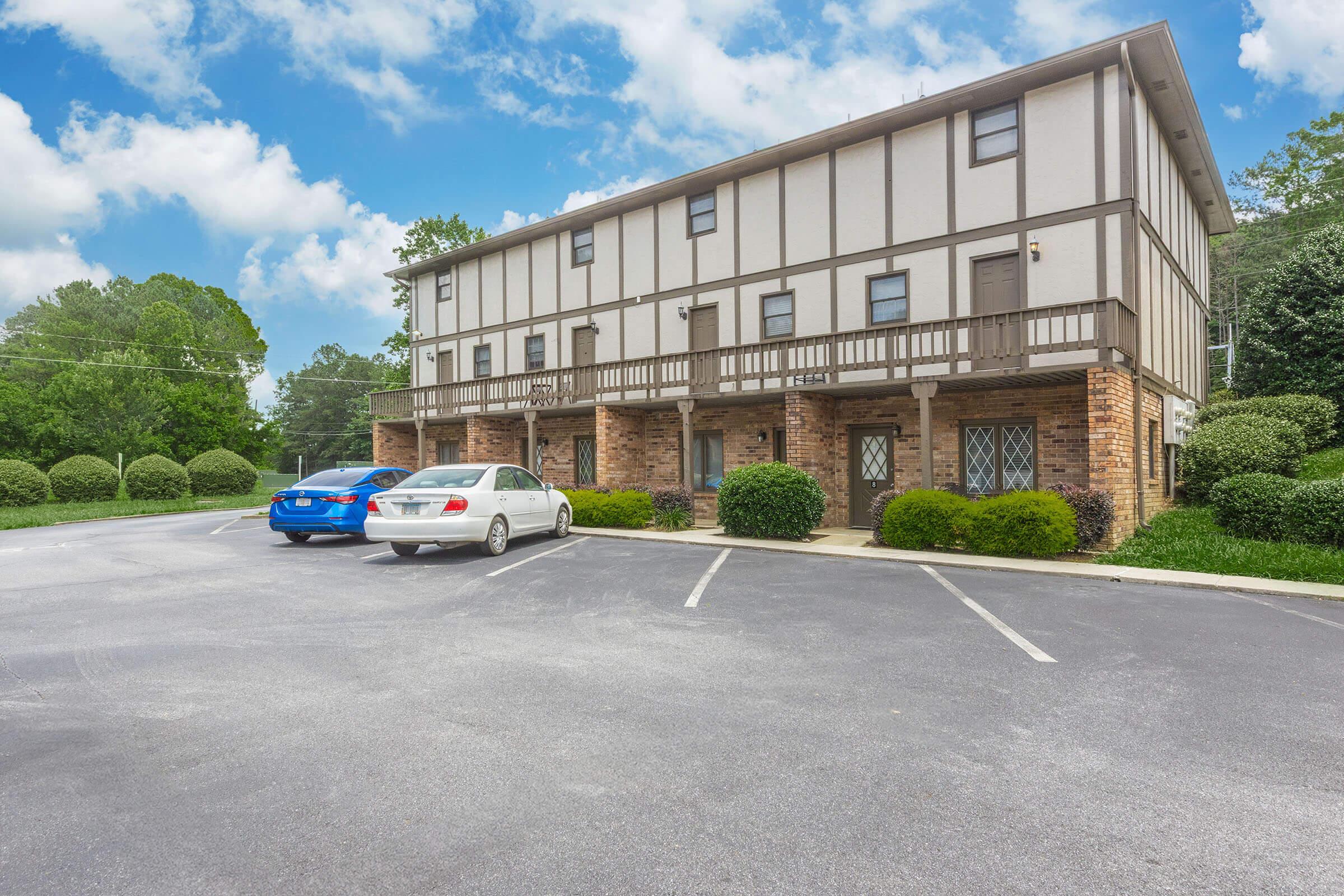A residential building with a Tudor-style exterior featuring brown wood accents and brick on the lower level. Two cars are parked in the foreground, with a landscaped area including round bushes. The sky is partly cloudy, and trees are visible in the background.