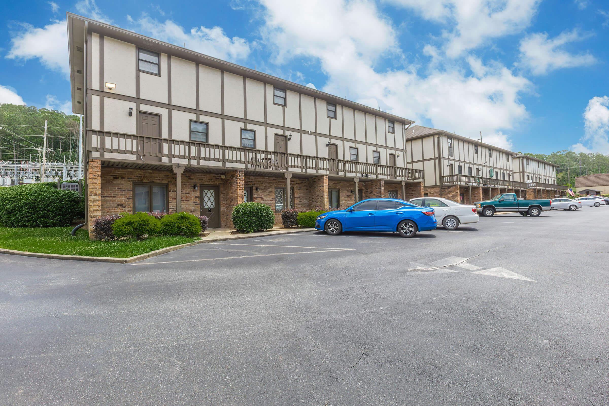Two multi-story apartment buildings with a modern architectural design. The foreground features a parking lot with several parked cars, including a blue vehicle. Lush greenery surrounds the buildings, and the sky is partly cloudy, creating a bright and inviting atmosphere.