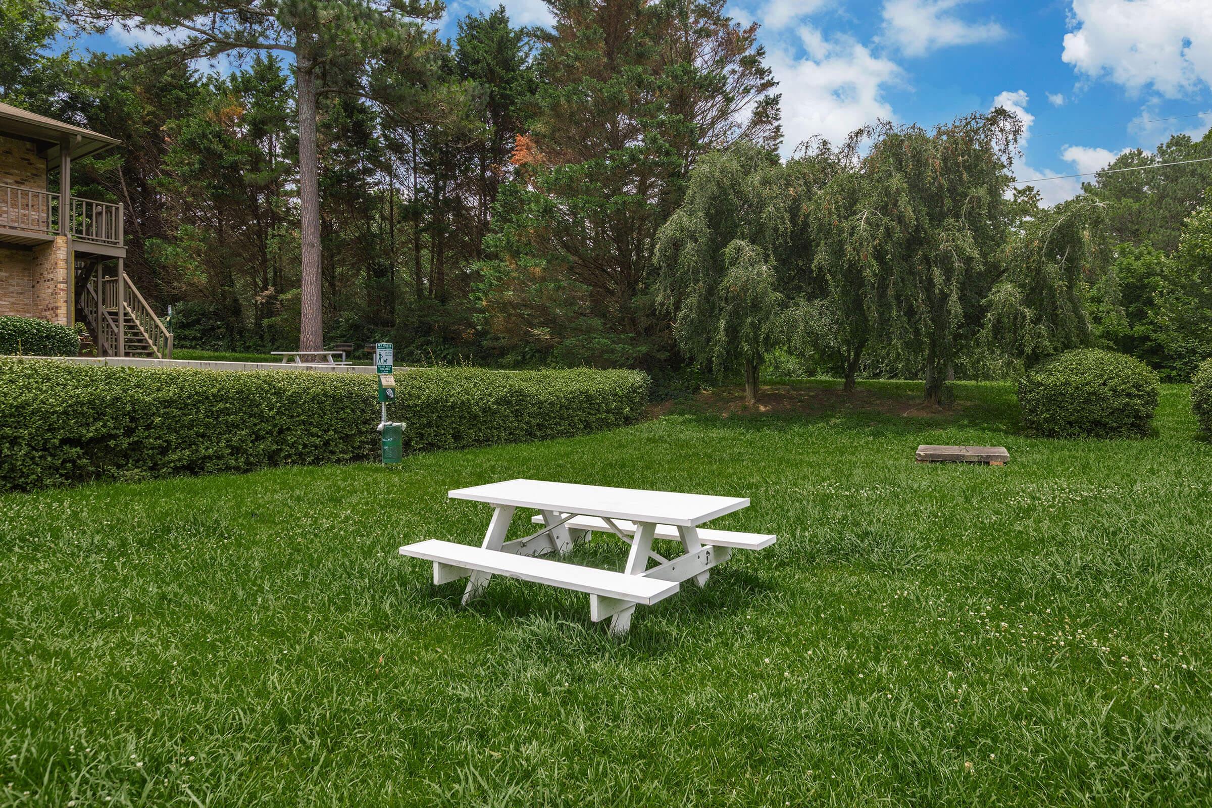 A picnic table in a grassy area surrounded by neatly trimmed bushes and trees, with a blue sky and a few clouds in the background. The setting is peaceful and inviting, ideal for outdoor gatherings or relaxation. A small fire pit is also visible in the background.
