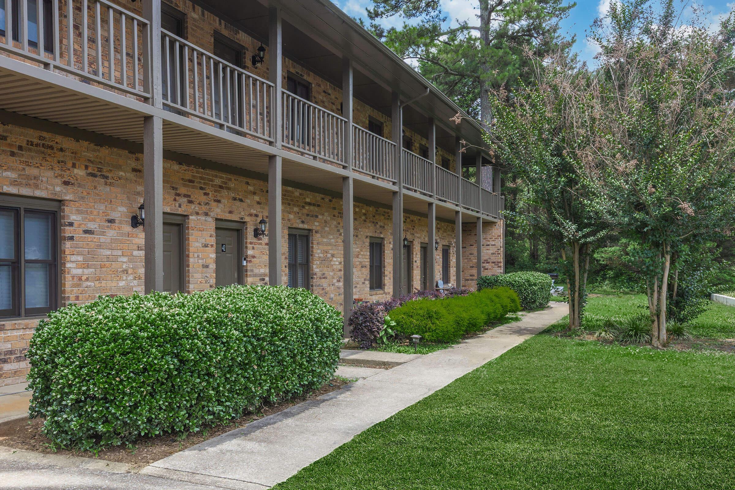 View of a two-story brick apartment building with a walkway leading to the entrance. The building features multiple balconies and neatly trimmed bushes along the path. Lush green grass and trees are visible in the surrounding area, creating a pleasant residential atmosphere.