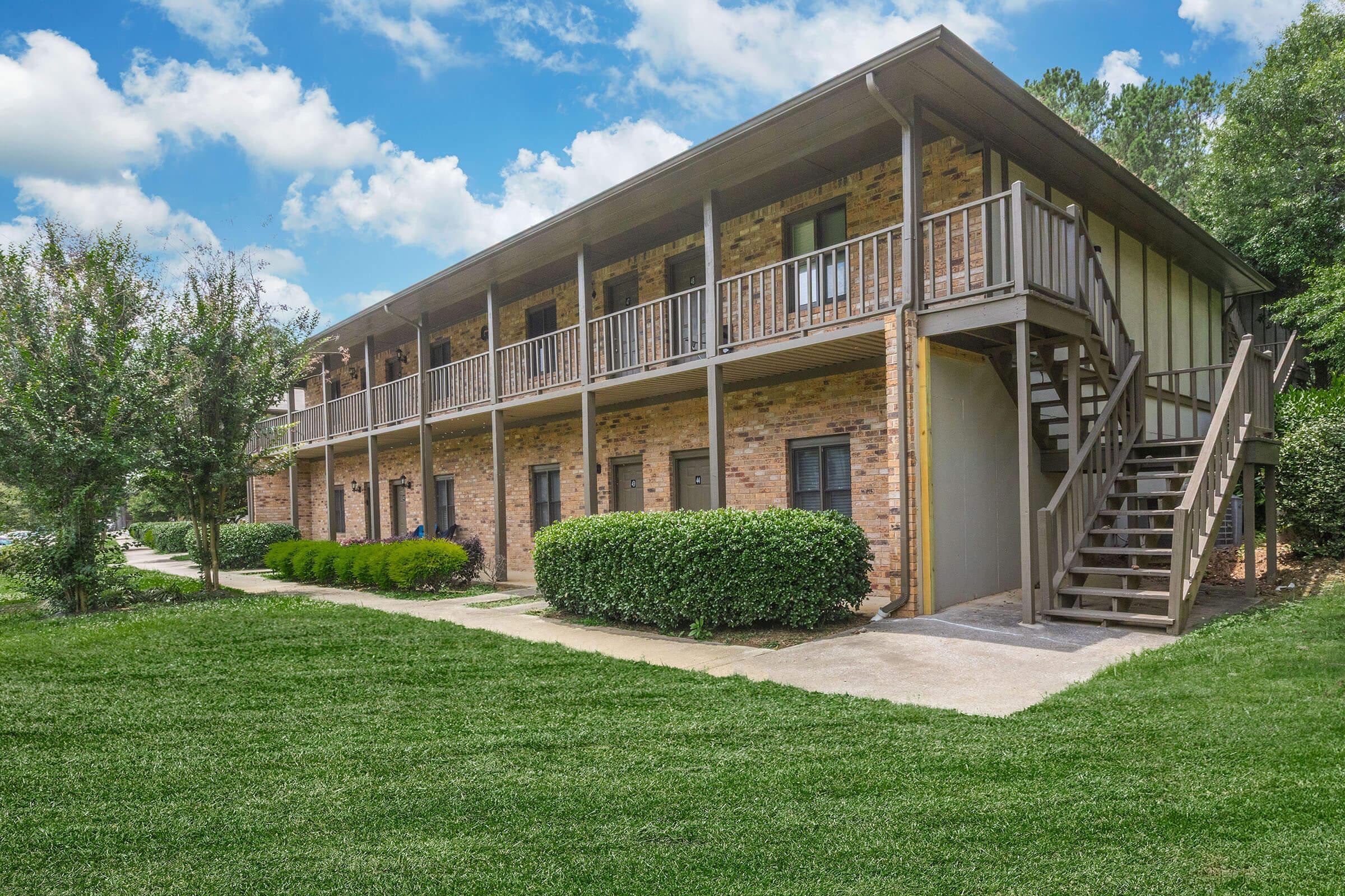 A multi-story apartment building with brick exterior and wooden balconies. The building has multiple units, each with a staircase leading to the second floor. Surrounding the building are well-maintained lawns and shrubbery, set against a backdrop of clear blue skies with a few clouds.