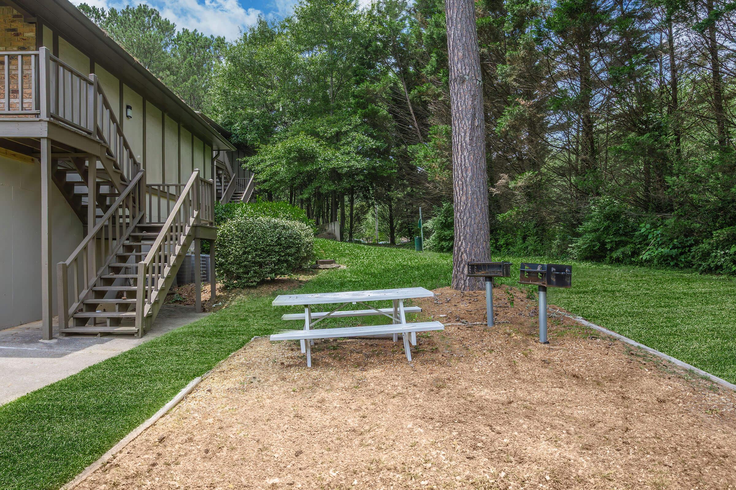 A picnic area featuring a white wooden table and two grills located near a building. Lush green grass surrounds the area, with trees in the background and steps leading up to an upper level of the building. The scene is bright and inviting, ideal for outdoor gatherings.