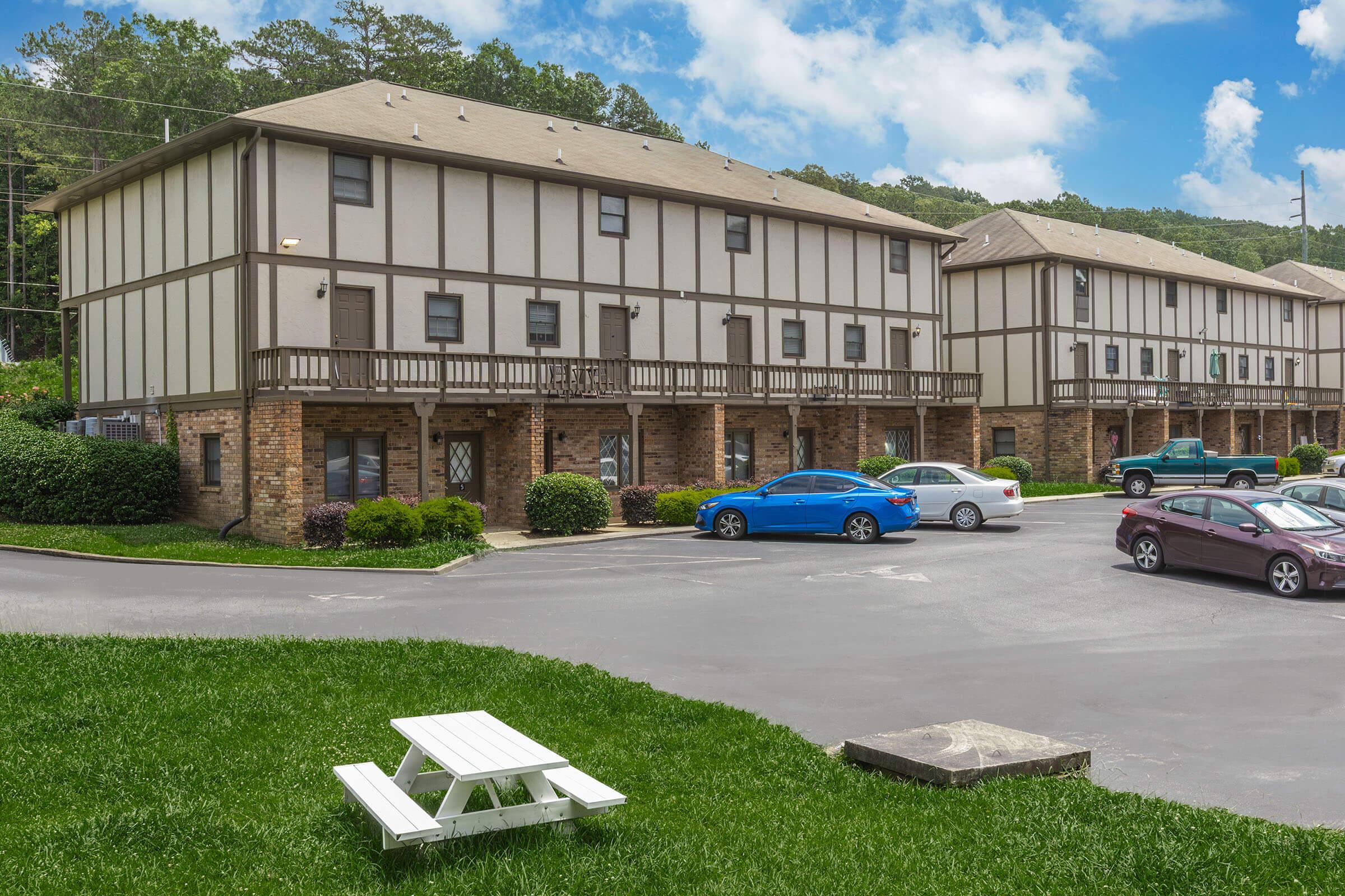 A row of two-story buildings with a Tudor architectural style, featuring white and brown exteriors. The foreground includes a grassy area with a picnic table, while a few cars are parked in the lot. Surrounding greenery and trees are visible under a blue sky with scattered clouds.