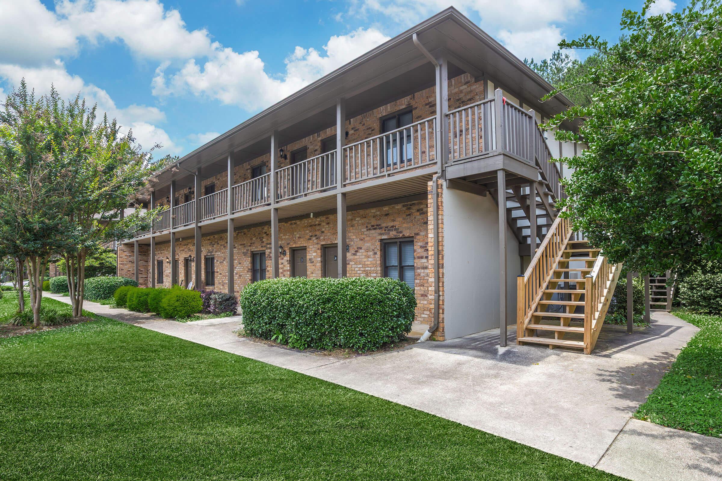 A multi-story brick apartment building with a wooden staircase on the side. Lush green shrubs and grass surround the property, and the sky is partly cloudy. The building features balconies on the upper level for each unit.