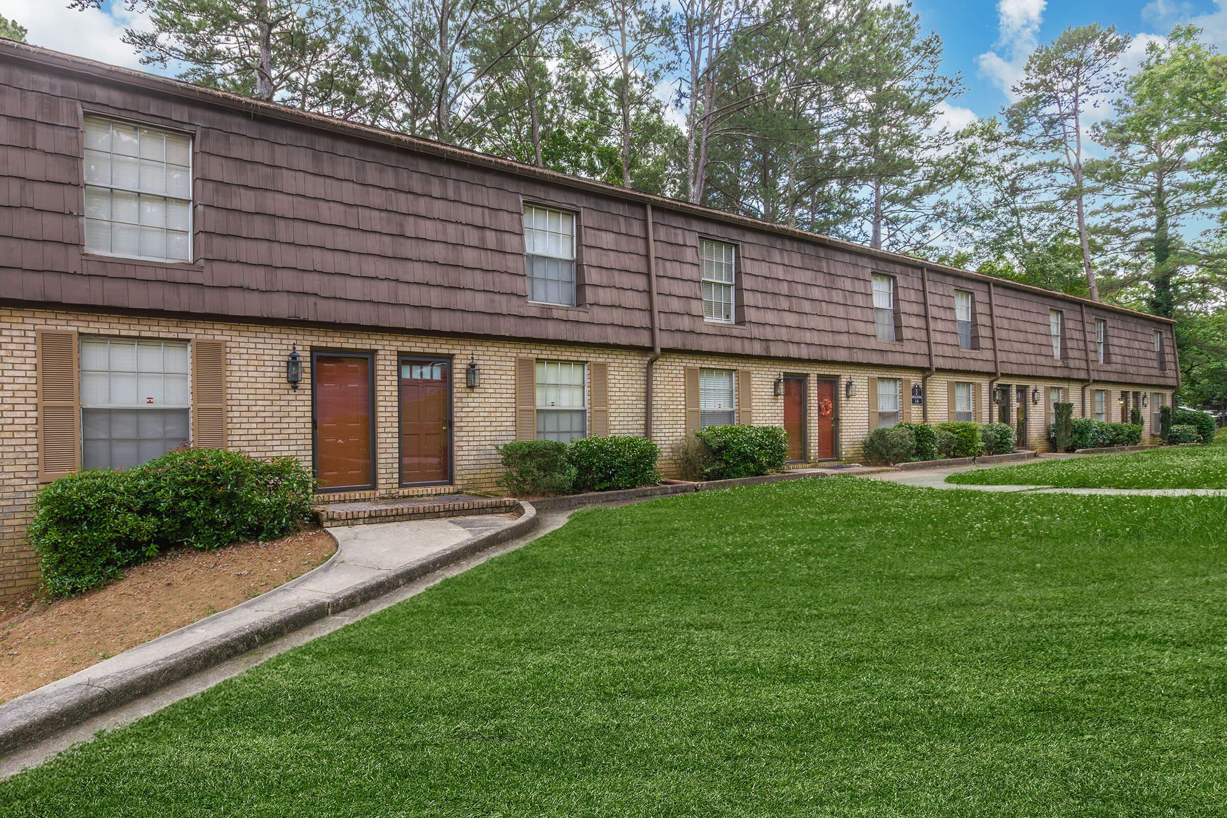 A row of two-story townhouses with brown shingle roofs and light brick exteriors. Each unit has a front door, with one door painted red. Green grass surrounds the buildings, and there are small shrubs planted along the walkway leading up to the doors. The scene is set against a backdrop of trees under a clear blue sky.