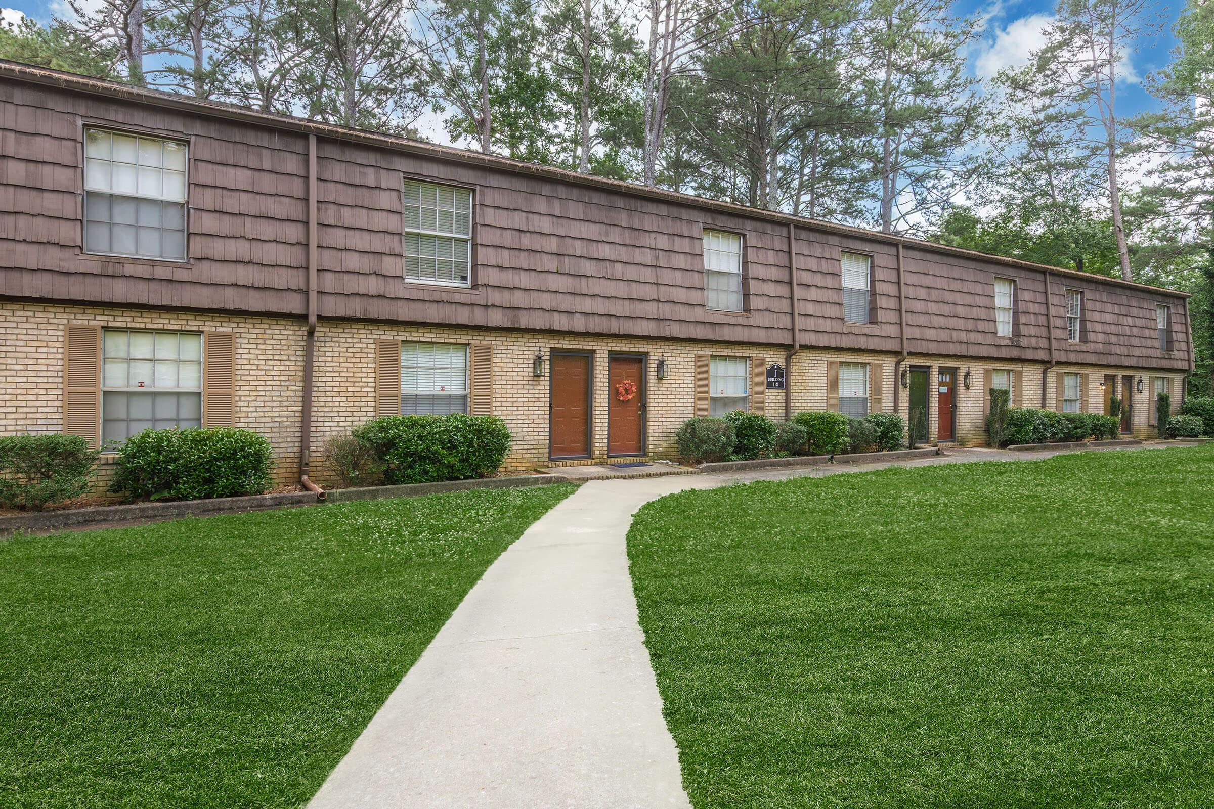 A row of brown, textured townhouses with wooden doors and wreaths, set against a backdrop of green grass and tall trees. A concrete pathway leads up to the front doors, and the sky is partly cloudy. The atmosphere is peaceful and inviting.