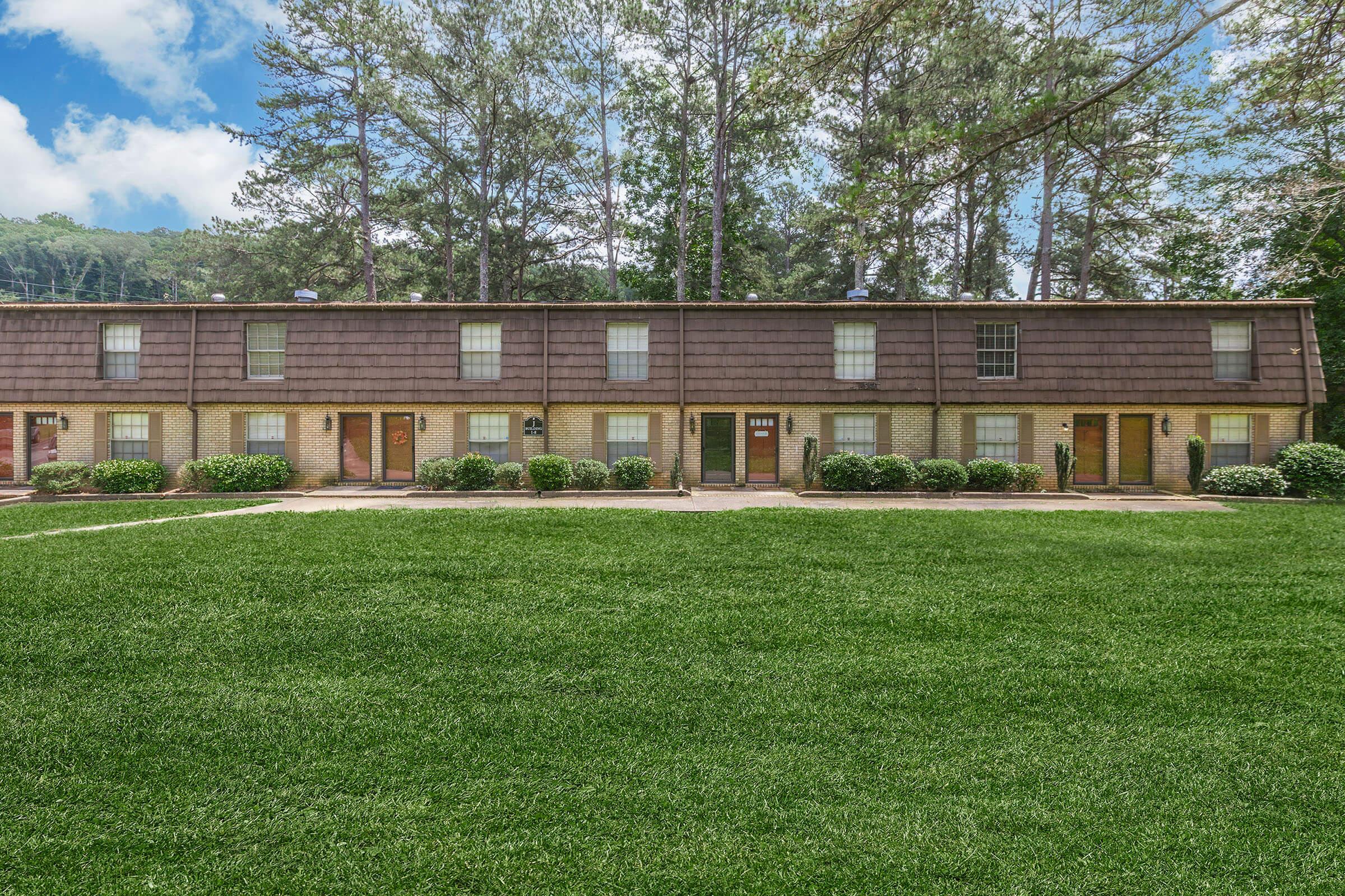 A low-rise apartment complex with a row of attached units featuring brown shingle roofs. The building is set in a grassy area with well-maintained bushes in front. Tall trees are in the background under a partly cloudy sky, suggesting a peaceful residential setting.