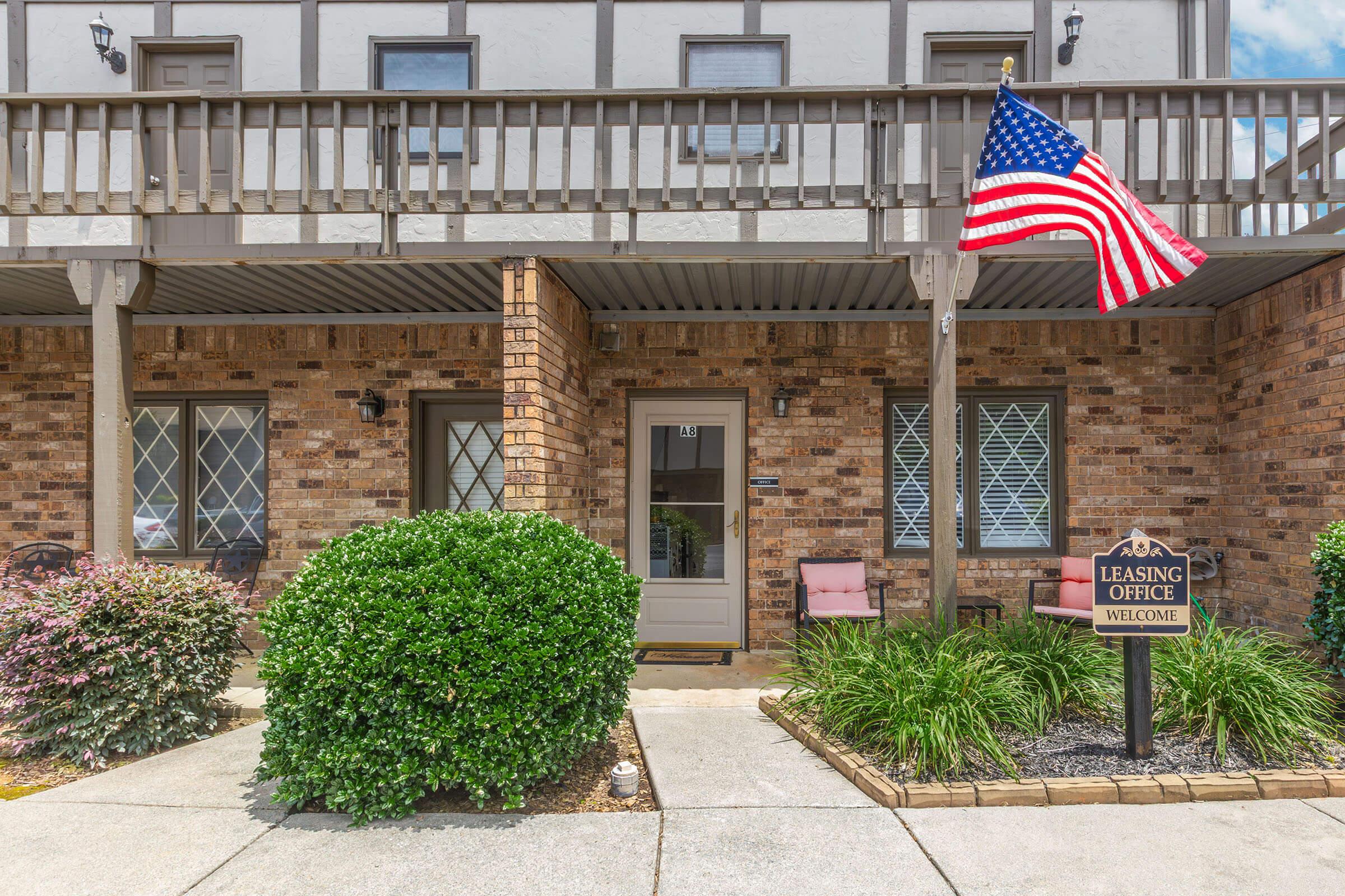 A leasing office entrance featuring a brick facade, a welcoming sign, and a neatly trimmed shrub in front. An American flag hangs on the balcony railing, with two chairs positioned near the door. The area is well-maintained, showcasing a welcoming atmosphere for potential tenants.