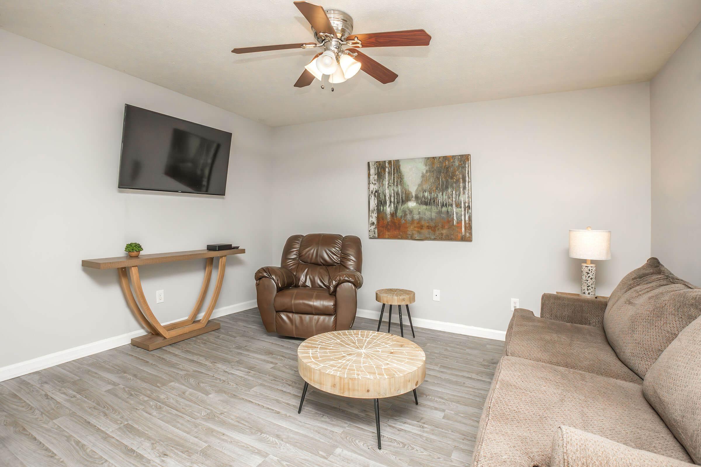 A cozy living room featuring a brown recliner chair, a light brown sofa, a wooden coffee table, and a side table with a lamp. A flat-screen TV is mounted on the wall, and a nature-themed artwork hangs nearby. The room has light gray walls and a ceiling fan, providing a comfortable and inviting atmosphere.
