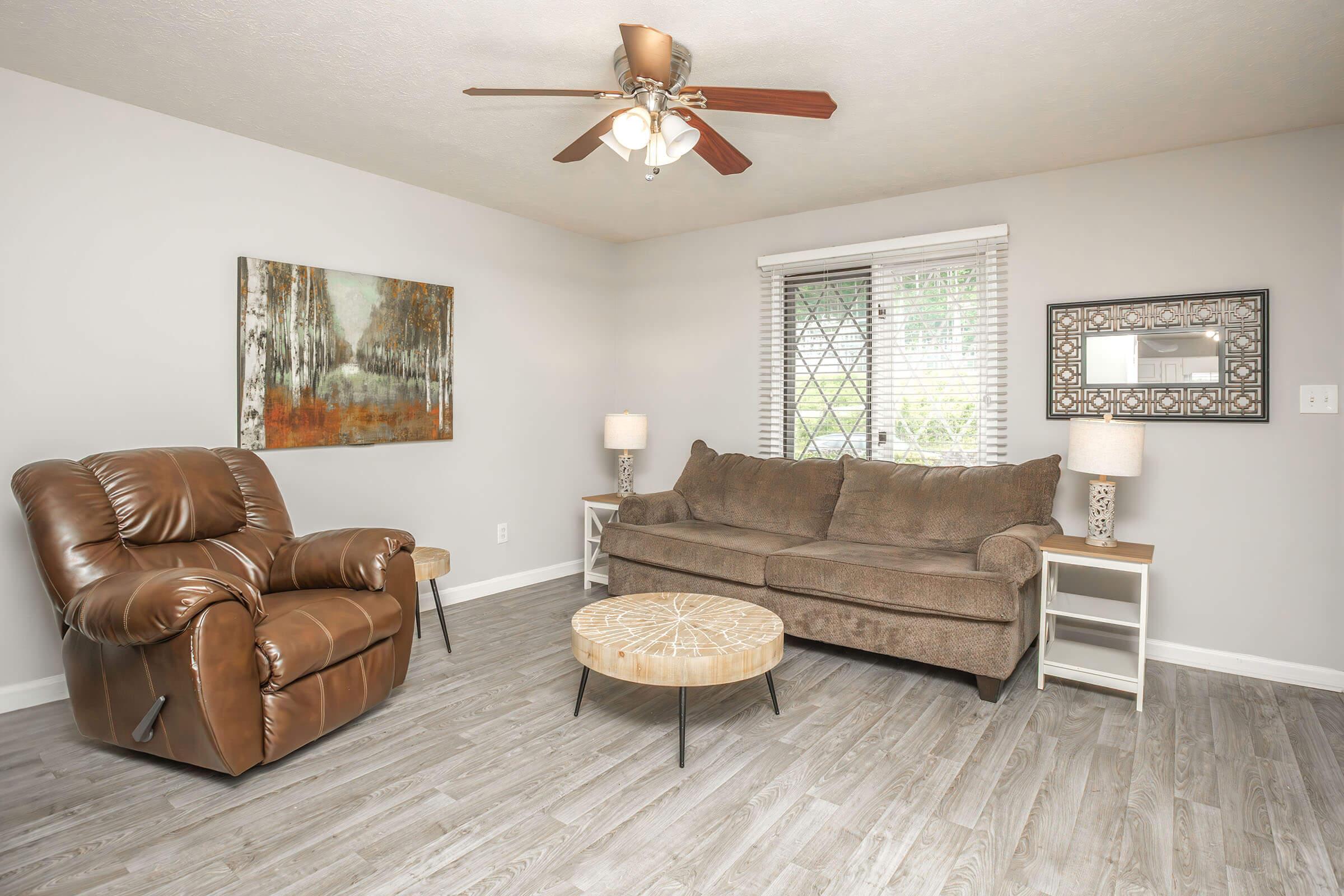 A cozy living room featuring a brown reclining chair, a brown sofa, a round coffee table, and two lamp stands. The walls are painted light gray, and a piece of artwork hangs on one wall near a window with blinds. The floor is light-colored wood laminate.
