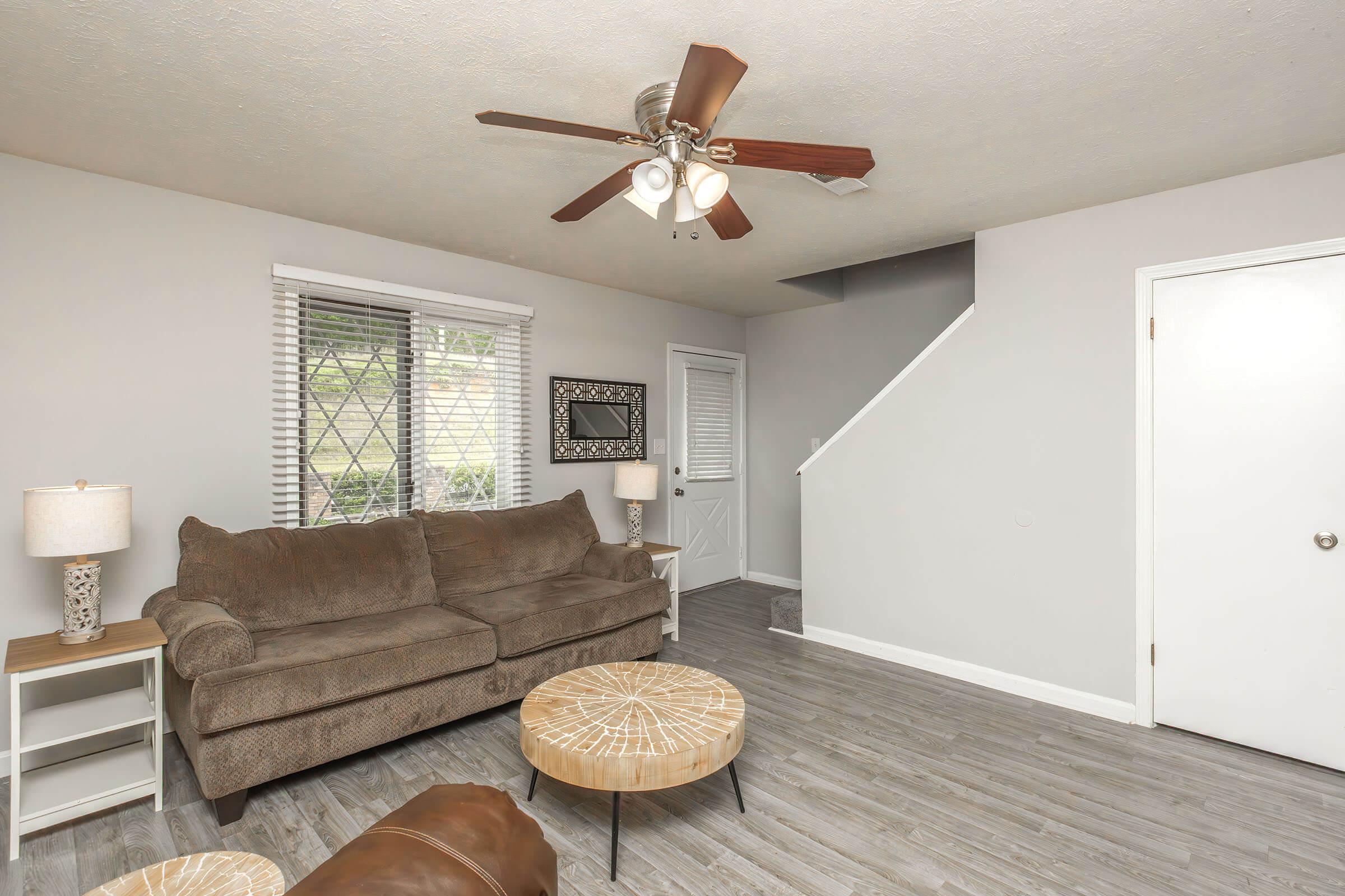A cozy living room featuring a brown sofa, two lamps on side tables, a round coffee table, and a ceiling fan. The room has light-colored walls, large windows with white blinds, and a staircase leading to an upper level. There's also a door visible, adding to the room's functionality.