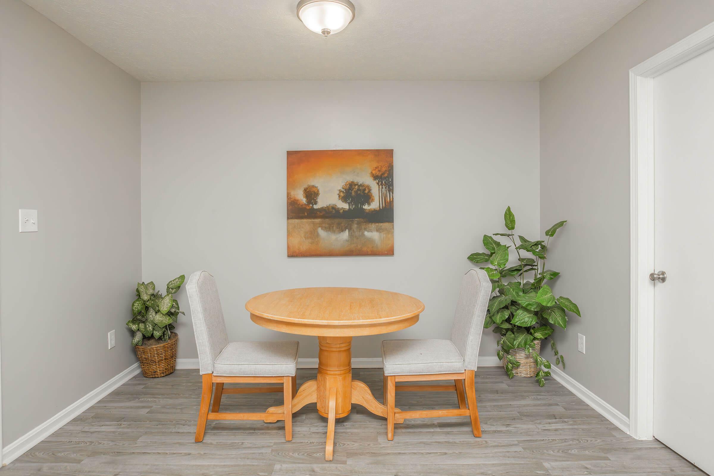 A small dining area featuring a round wooden table with two light gray chairs. The wall is painted in a soft gray color, and there is a landscape painting depicting trees and a lake. A potted plant adds a touch of greenery to the corner, enhancing the cozy atmosphere of the space.