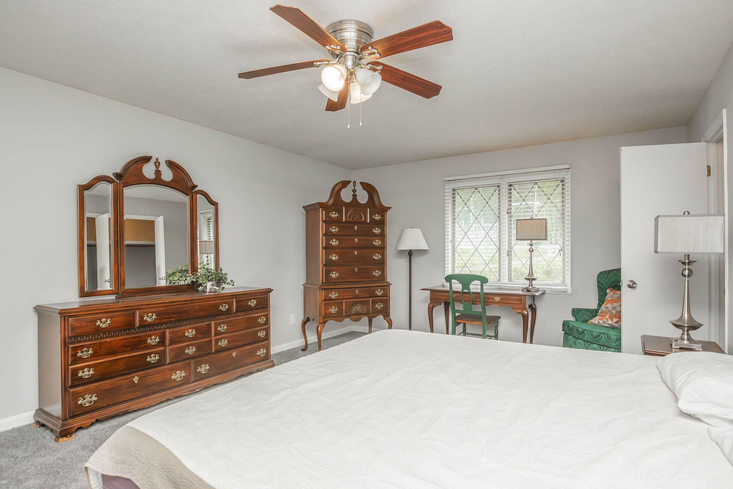A cozy bedroom featuring a large bed with a white quilt, a wooden dresser with decorative handles, a matching tall chest of drawers, and a small desk with a green chair. The room has a ceiling fan and a window with sheer curtains, providing natural light. A green armchair is positioned by the window.