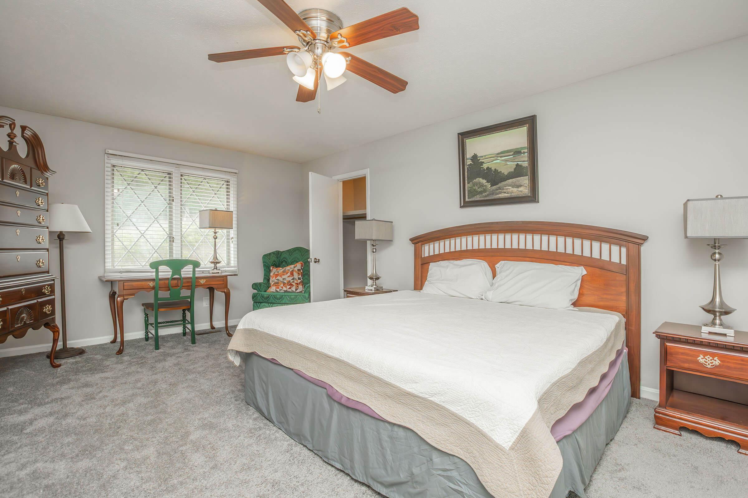 A cozy bedroom featuring a large wooden bed with white bedding, a classic dresser, a green chair, and lamps on bedside tables. The walls are painted in a light color, and there's a ceiling fan and artwork on the wall. Natural light streams through the window with a view outside.