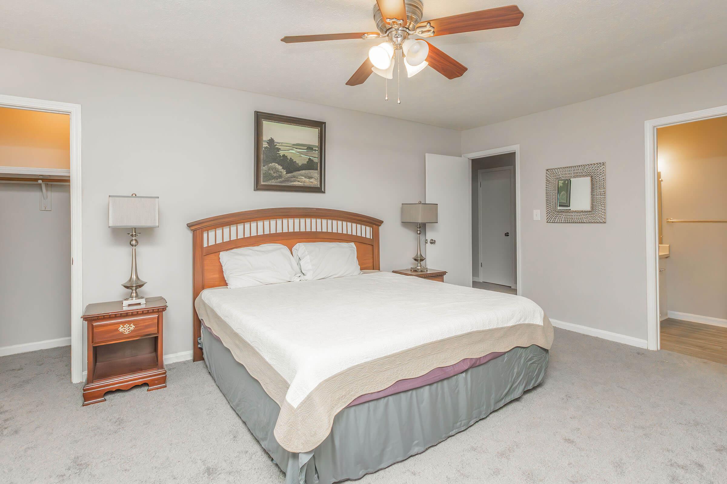 A well-decorated bedroom featuring a wooden bed frame with a light-colored bedding set, two bedside tables with lamps, and a ceiling fan. The walls are painted in a soft gray, and there are two doors leading to closets and a bathroom. A landscape painting is hung above the bed.