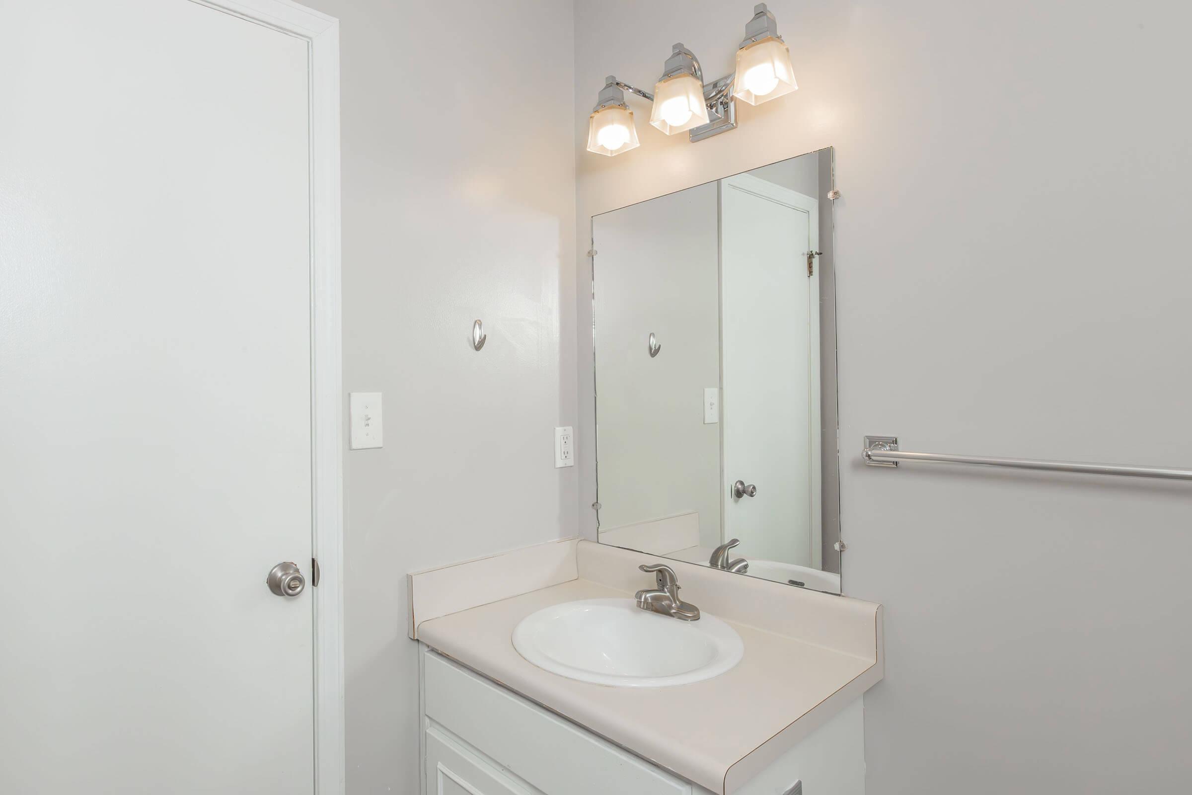 A small bathroom featuring a white sink with a polished faucet, a large mirror above the sink, and three light fixtures above it. The walls are painted light gray, and there is a white door leading to another room. A towel bar is mounted beside the mirror, completing the minimalist design.