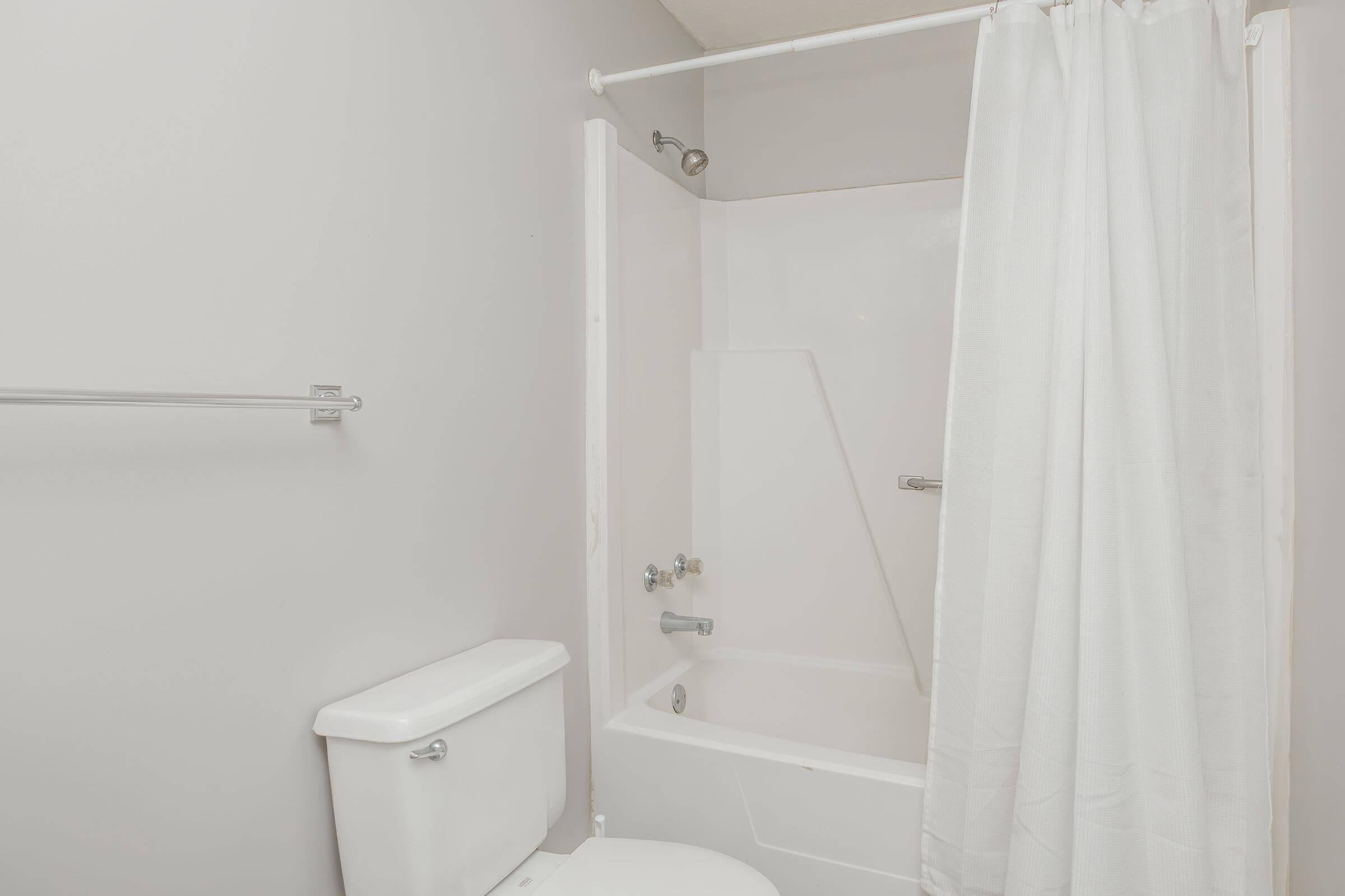 A small, simple bathroom featuring a shower-tub combination with a white curtain, a toilet, and a towel rack. The walls are painted light gray, contributing to a clean and minimalistic aesthetic.