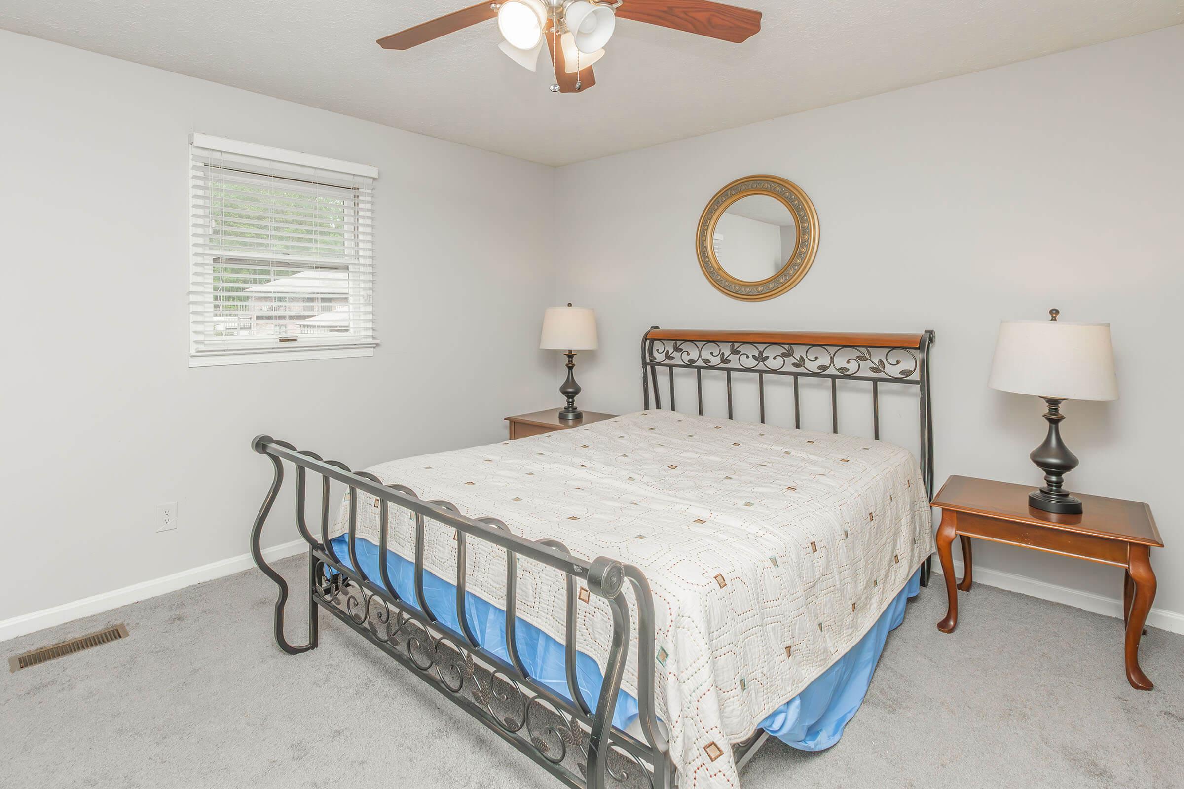 A cozy bedroom featuring a wrought iron bed with a light-colored quilt, two wooden nightstands with lamps, a round mirror on the wall, and a window with white blinds. The walls are painted light gray, and the floor is covered with carpet, creating a warm and inviting atmosphere.