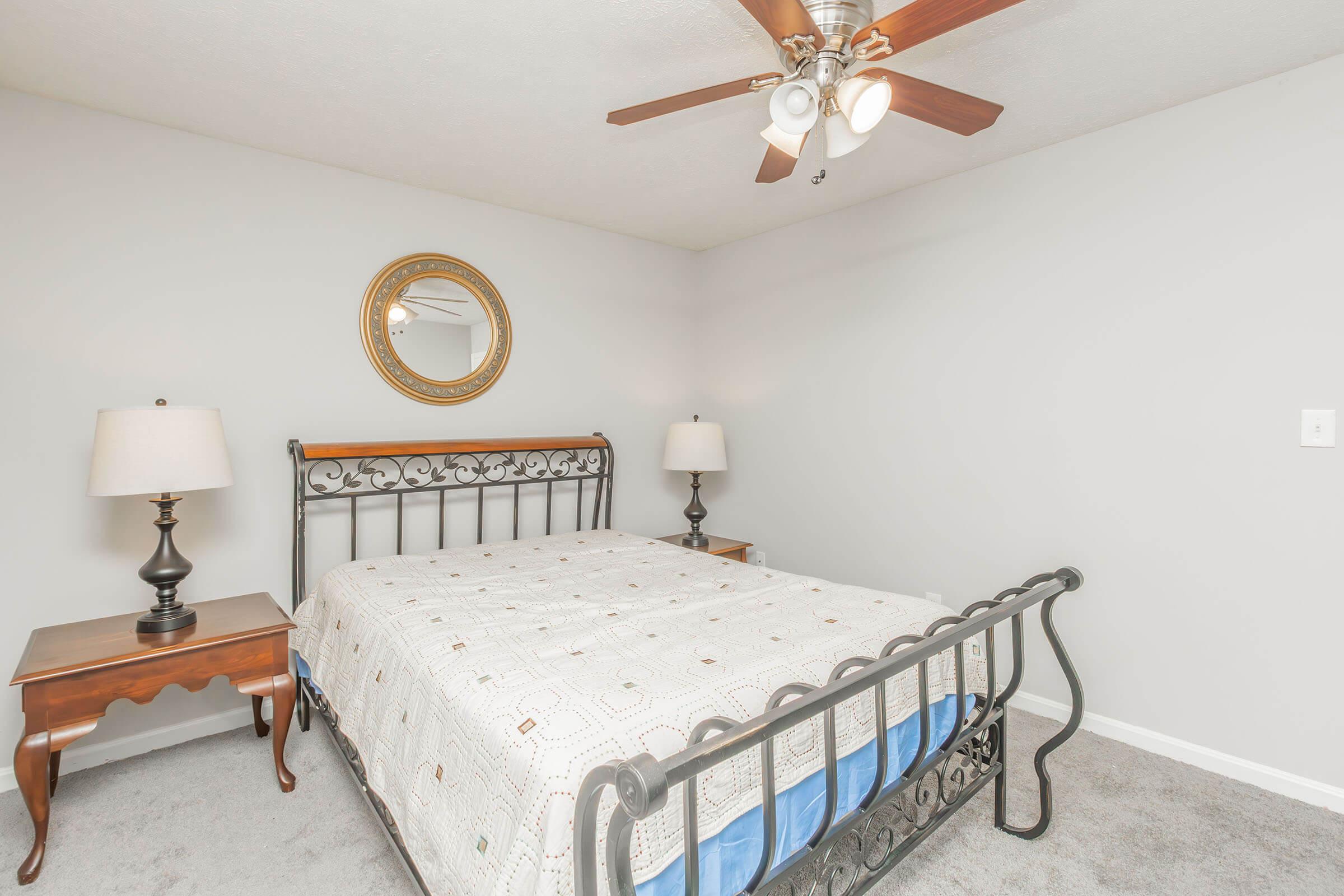A cozy bedroom featuring a metal bed frame with a patterned bedspread, two bedside tables with lamps, and a round mirror on the wall. The room has light gray walls and a ceiling fan, creating a comfortable and inviting atmosphere.