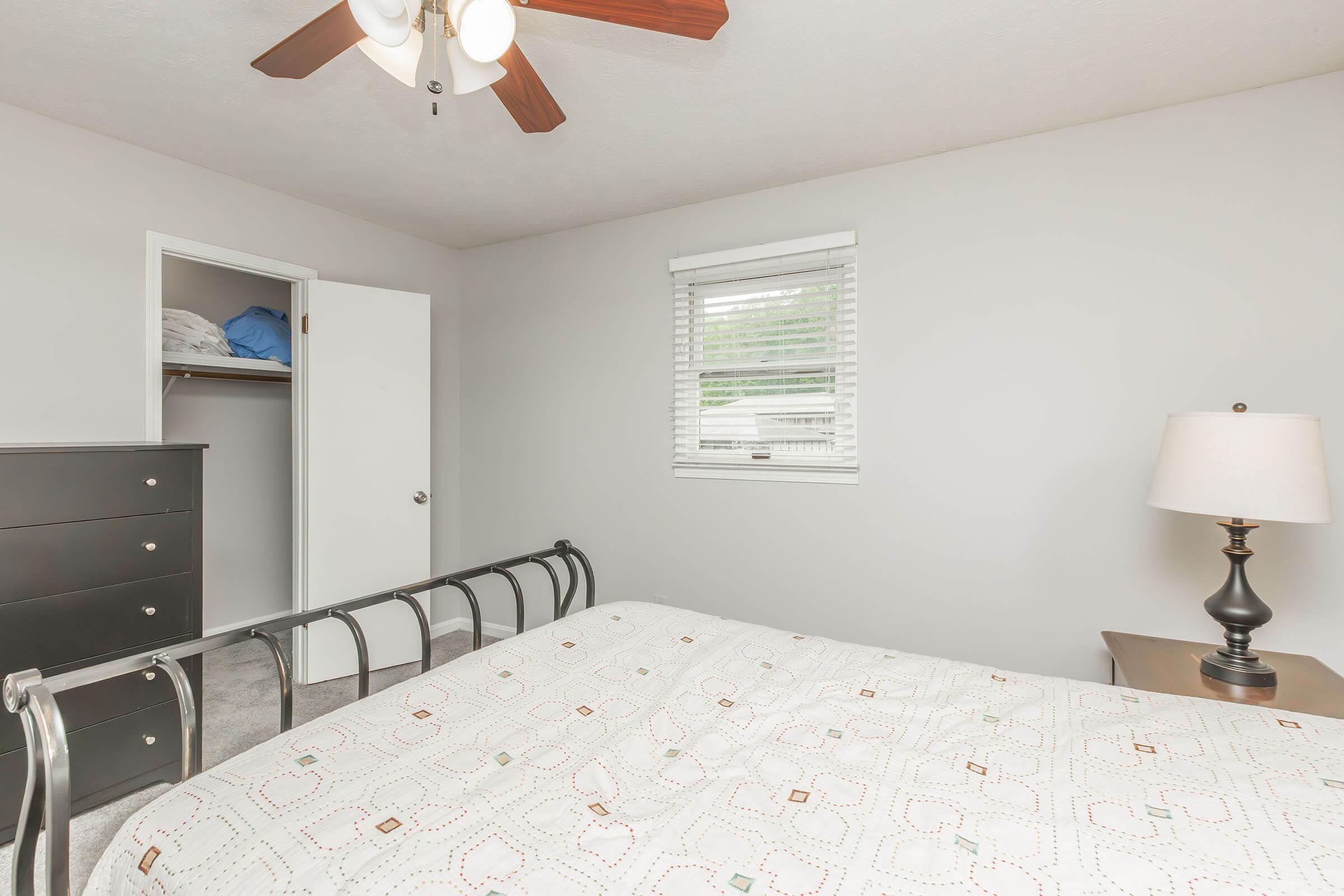 A cozy bedroom featuring a bed with a patterned comforter, a bedside lamp on a wooden nightstand, and a chest of drawers. A closet with blue items is visible in the background, along with a window allowing natural light to enter the room. The walls are painted a light gray, enhancing the room's brightness.