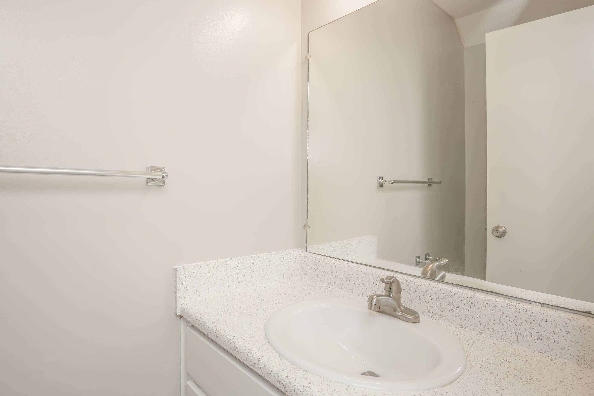 A clean, modern bathroom featuring a white sink with a chrome faucet, a large mirror above the sink, a simple countertop, and a towel rack on the wall. A light-colored door is visible in the background, giving the space a bright and airy feel.