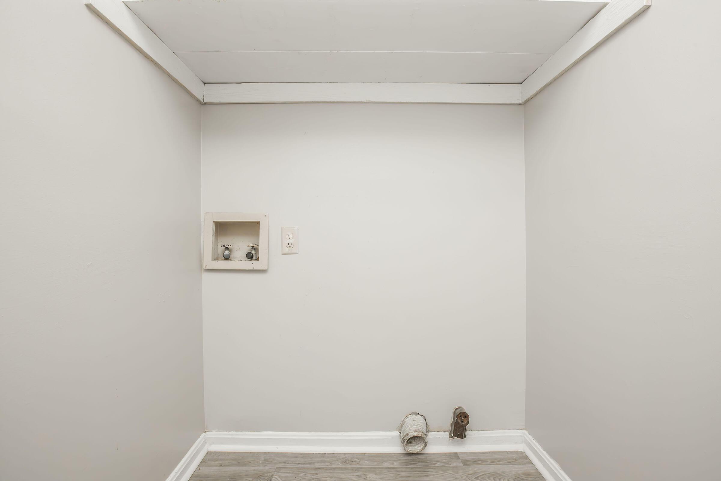 A minimalist room with light gray walls and a wooden floor. One corner features a small, recessed shelf with two decorative objects, while on the floor beside it, there’s a rolled-up piece of fabric and another decorative item. The overall atmosphere is simple and sparse.