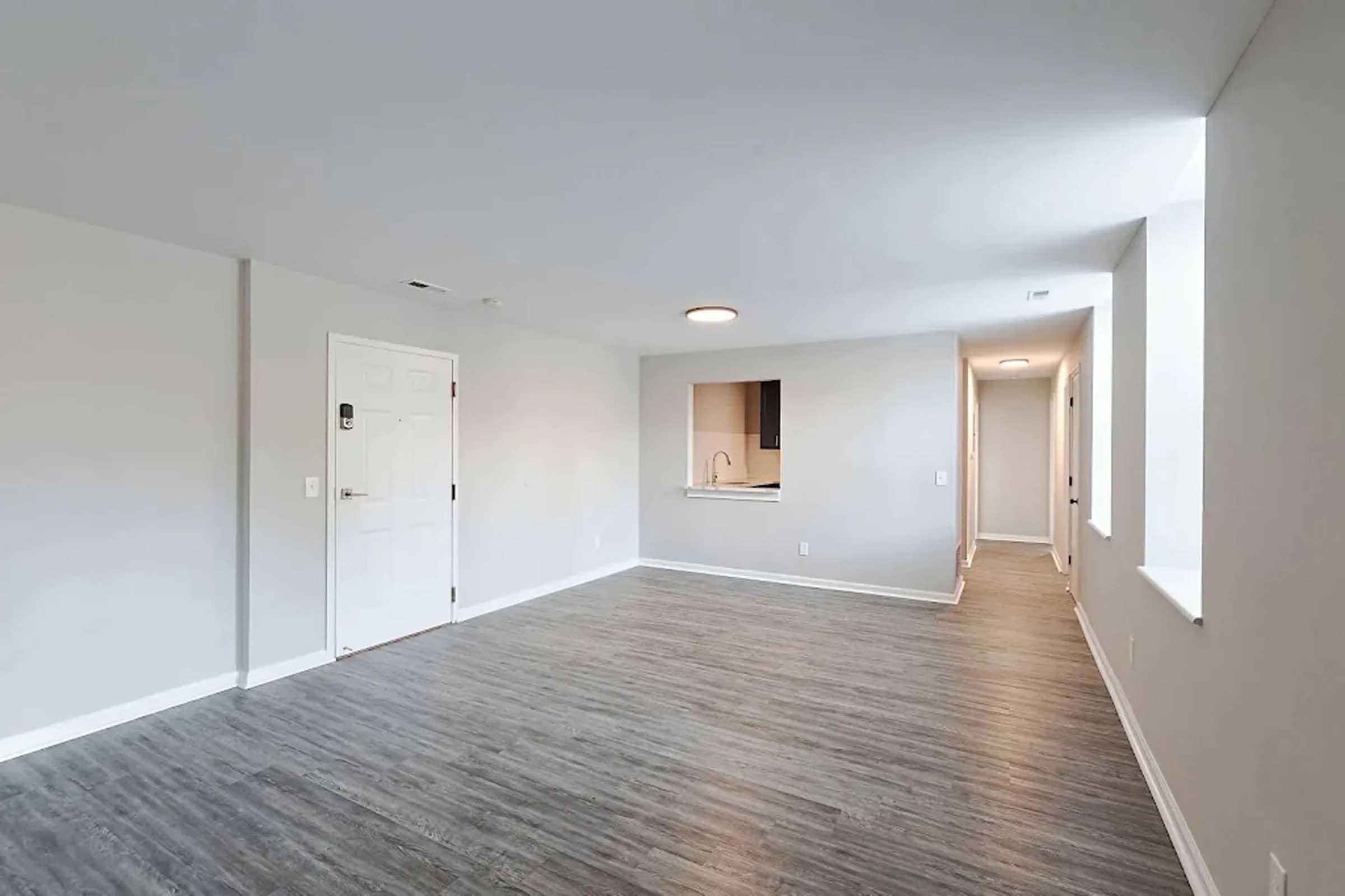 A spacious, empty room with light gray walls and laminate flooring. A white door is visible on the left, leading to an entrance. To the right, a doorway leads to a hallway. A large window allows natural light, and there's an open area in the wall that likely connects to a kitchen space.