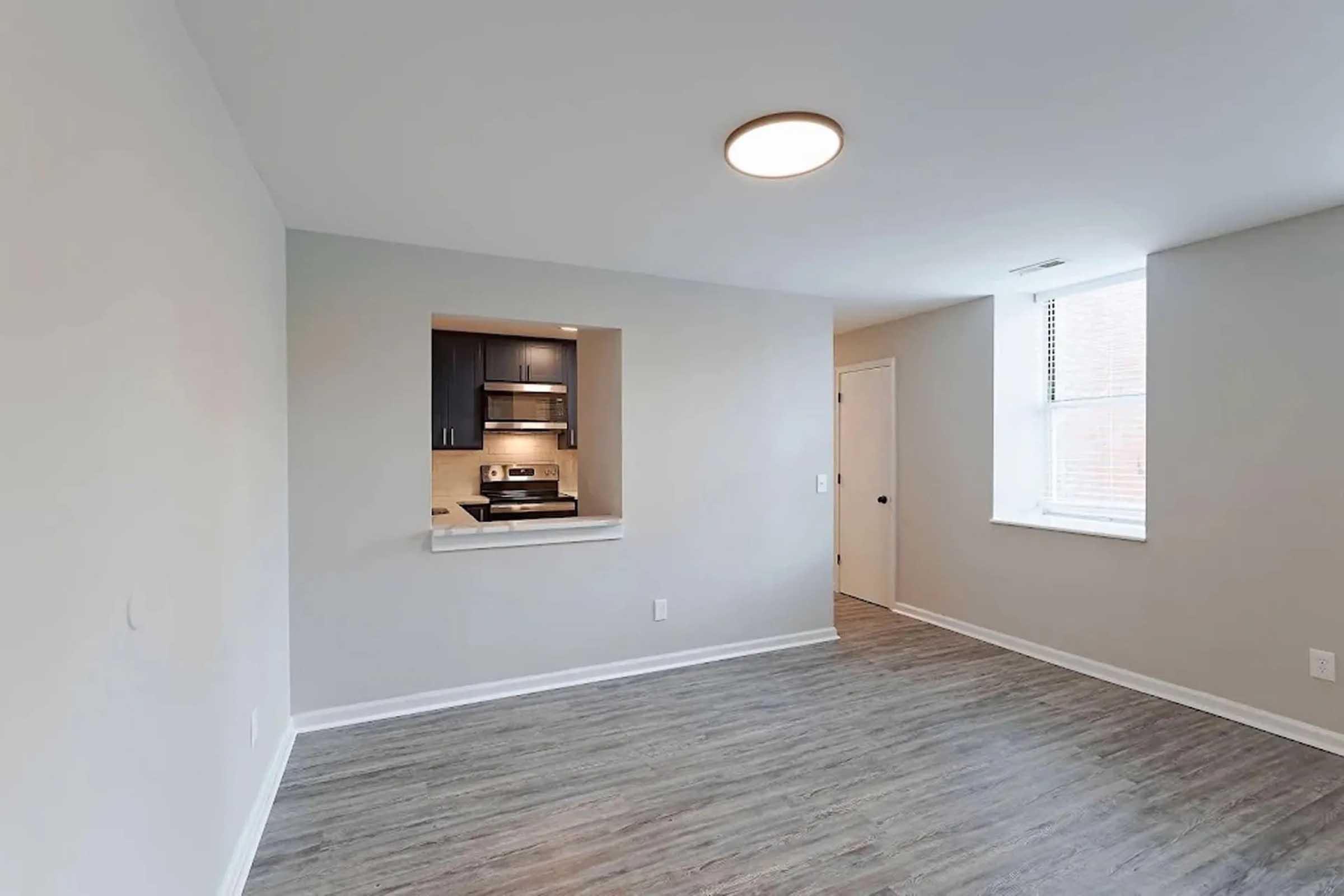 A bright, modern apartment interior featuring light gray walls and wood-like flooring. The space includes an open kitchen with dark cabinetry visible through a cut-out, a window providing natural light, and a door leading to another room. The overall atmosphere is spacious and inviting.