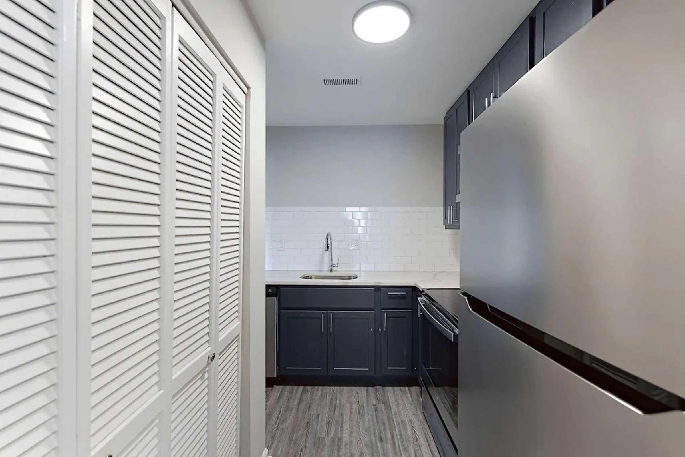 A modern kitchen featuring dark cabinetry, a stainless steel refrigerator, and a white tiled backsplash. The space includes a sink and is well-lit with a ceiling fixture. There are closed louvered doors on the left, providing storage or access to another area.