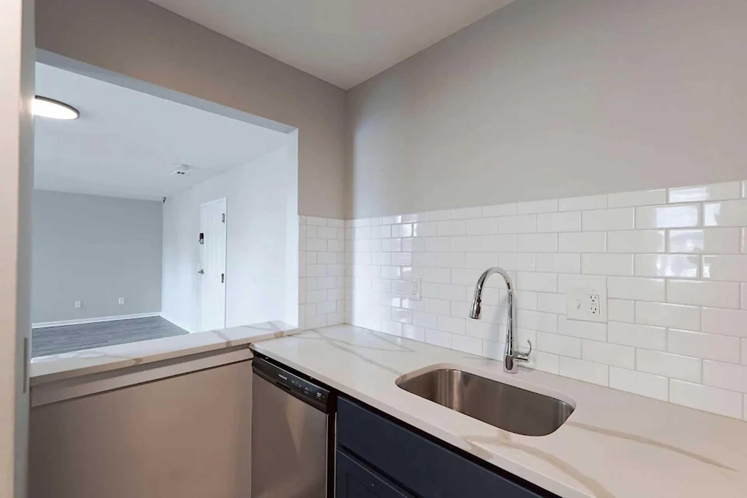 A modern kitchen scene featuring a stainless steel sink with a pull-down faucet, surrounded by white subway tile backsplash. A dishwasher is adjacent to the sink, and an open archway leads to a bright, empty living area with light gray walls and carpet.