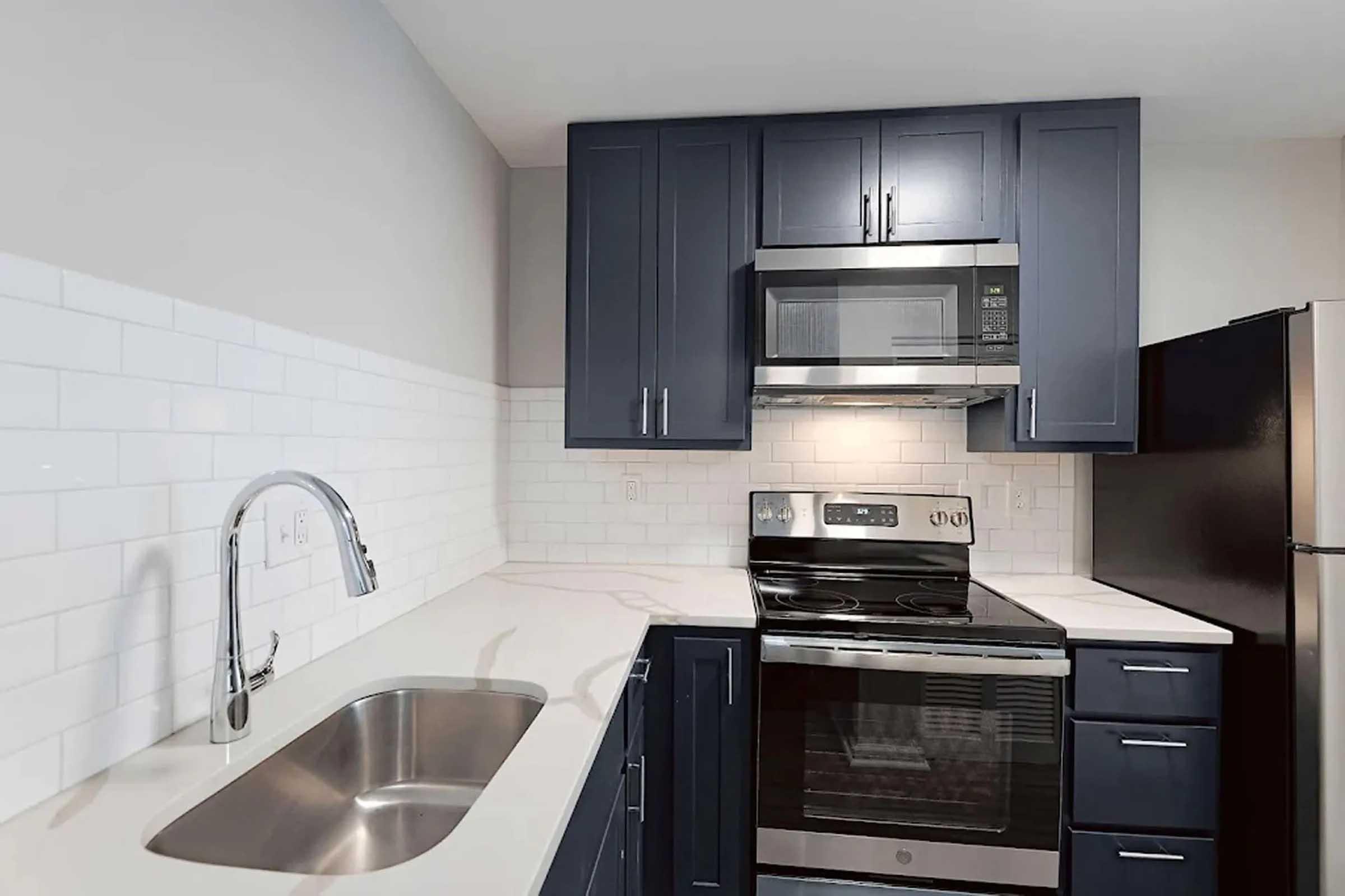 Modern kitchen featuring dark blue cabinetry, stainless steel appliances including a microwave and oven, a large sink with a sleek faucet, and white subway tile backsplash. The countertops are light-colored, complementing the overall contemporary design.