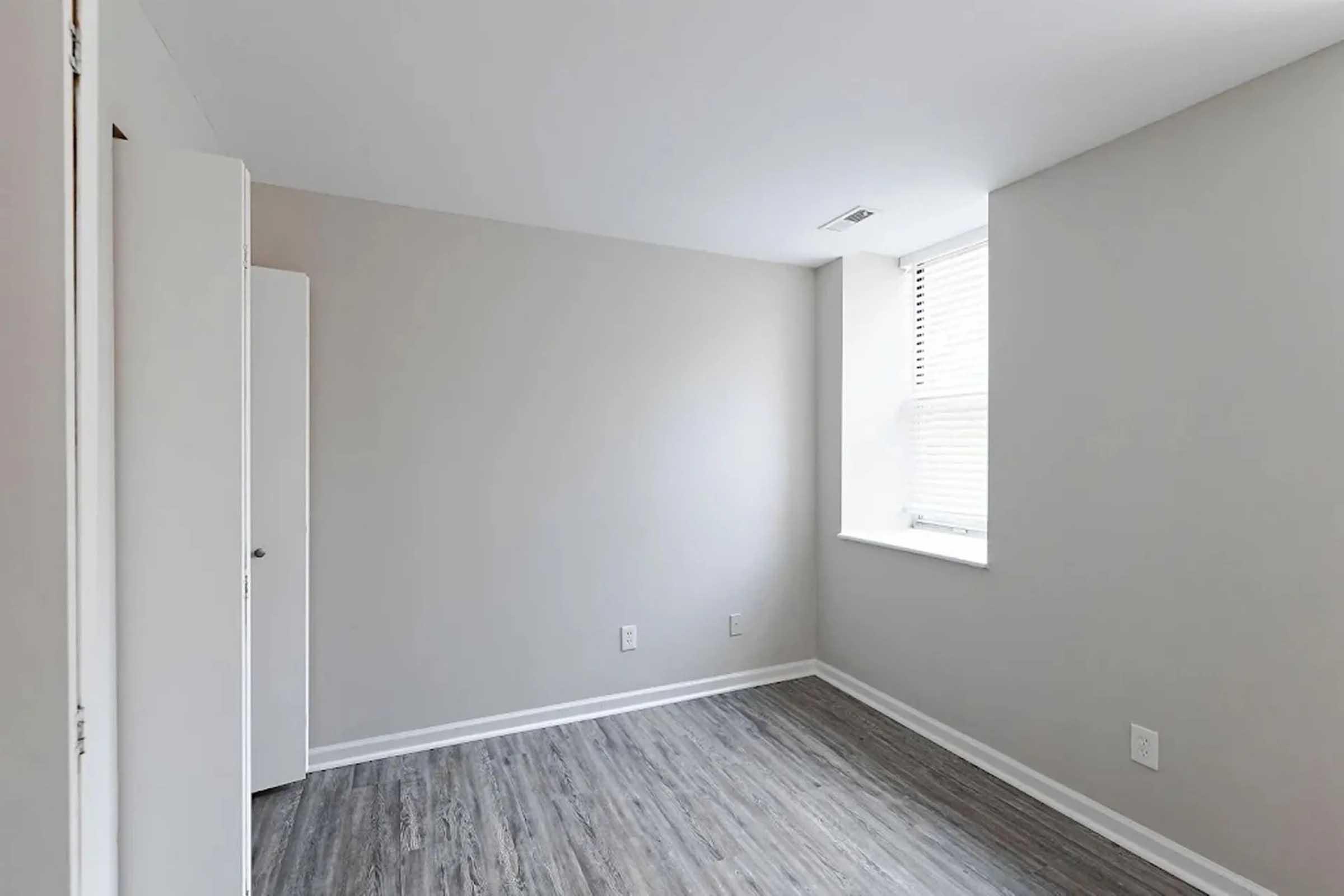 A vacant room with light gray walls and a wooden floor. It features a window on one wall allowing natural light, and a white door is partially visible to the left. The space is minimal and uncluttered, creating an open, airy feel.