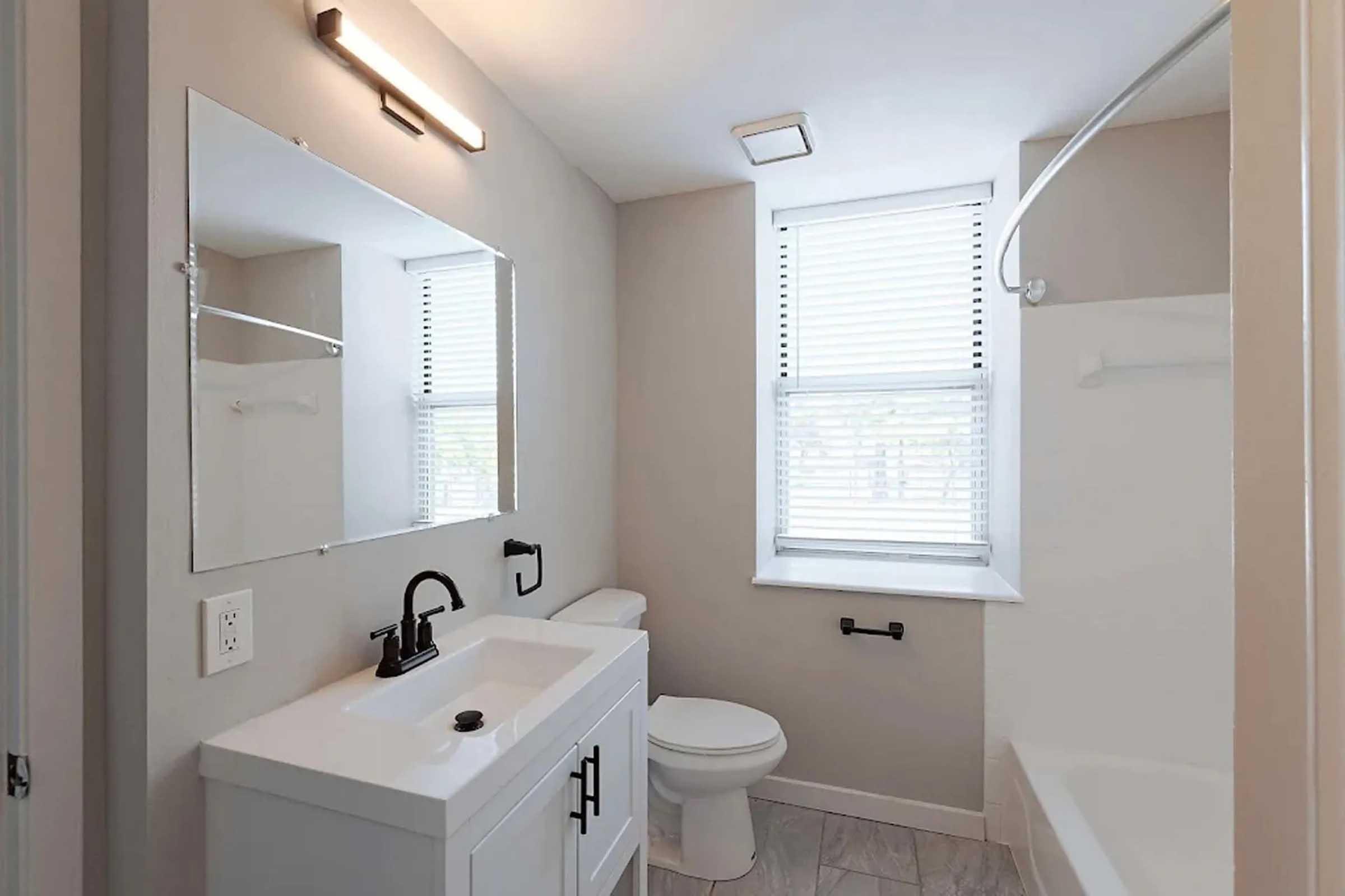 A modern bathroom featuring a white vanity with a black faucet, a large mirror above the sink, and a white toilet. There's a bathtub with a shower rod, and the space is illuminated by a ceiling light and natural light streaming through two windows with blinds. The walls are painted a neutral color.