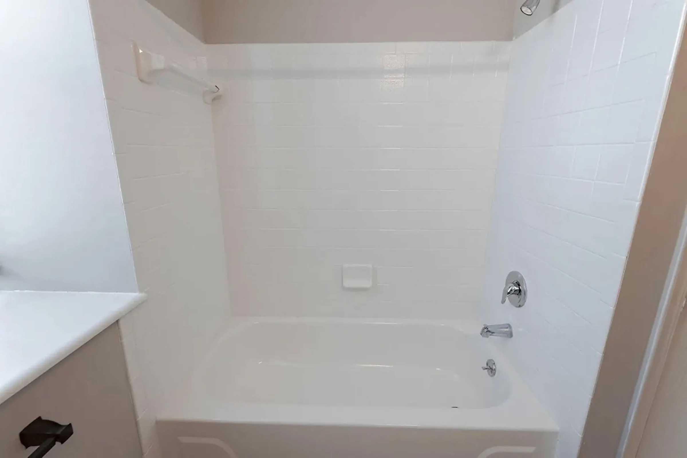 A clean, white tiled bathroom with a bathtub and shower combination. The walls are covered in white tiles, and there is a single faucet and showerhead. The space is well-lit and minimalist, featuring a simple design with no additional decor.